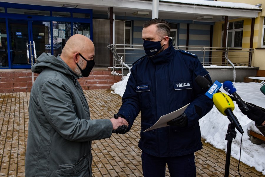 Komendant miejskiej policji, który w ręku trzyma list z podziękowaniami gratuluje mężczyźnie, który jest ubrany w szarą kurtkę