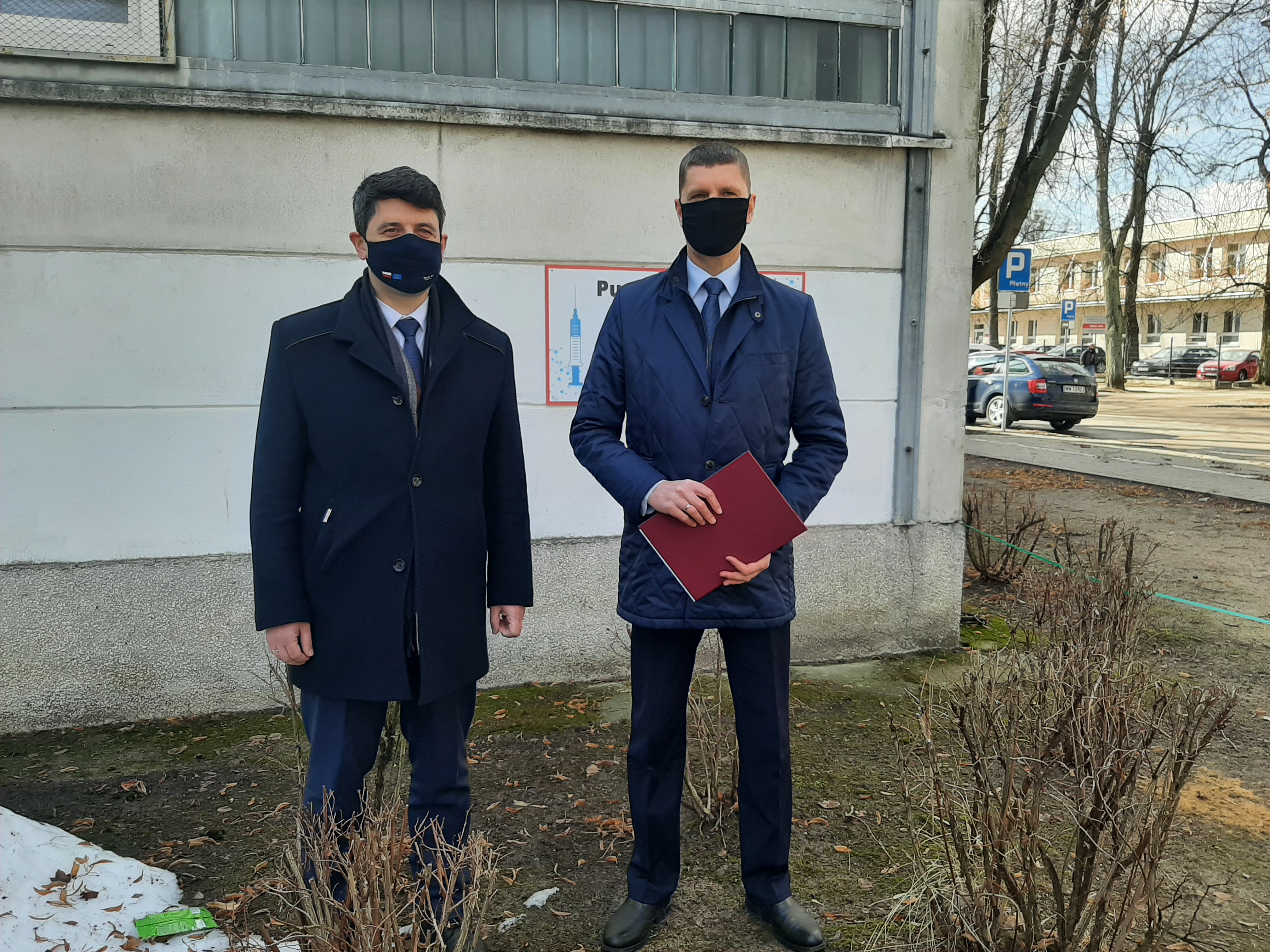 Poseł Mariusz Gromko, Dariusz Piontkowski stoją na tle betonowej ściany. Mężczyźni są ubrani w granatowe garnitury i płaszcze