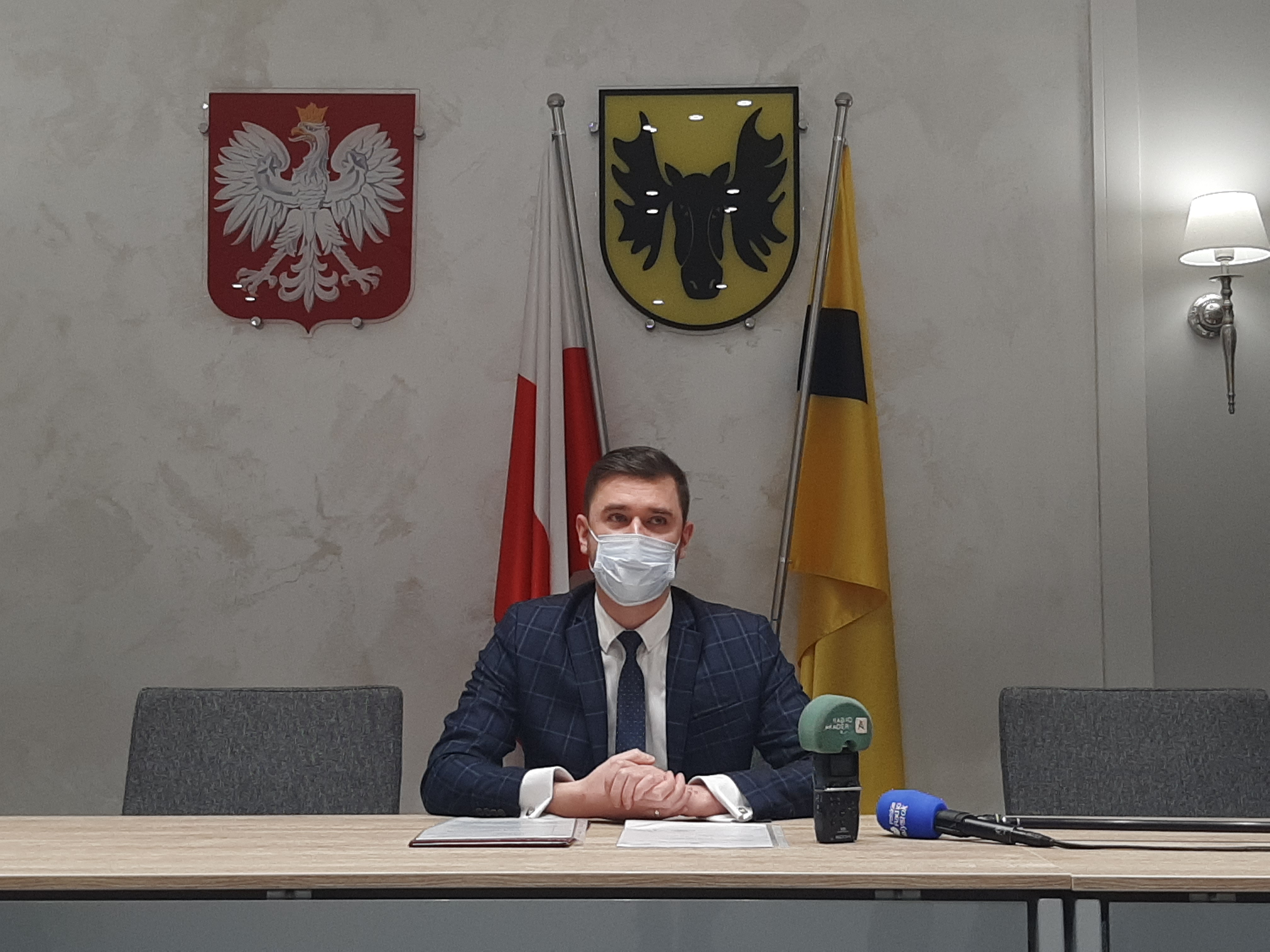 Mężczyzna w maseczce chirurgicznej siedzi przy stole. Za nim, po lewej stronie widoczne jest godło Polski, obok, po prawej stronie wisi herb Wasilkowa. Niżej stoją flagi, kolejno, Polski i Wasilkowa. Przy prawej krawędzi zdjęcia widoczna jest zapalona lampka