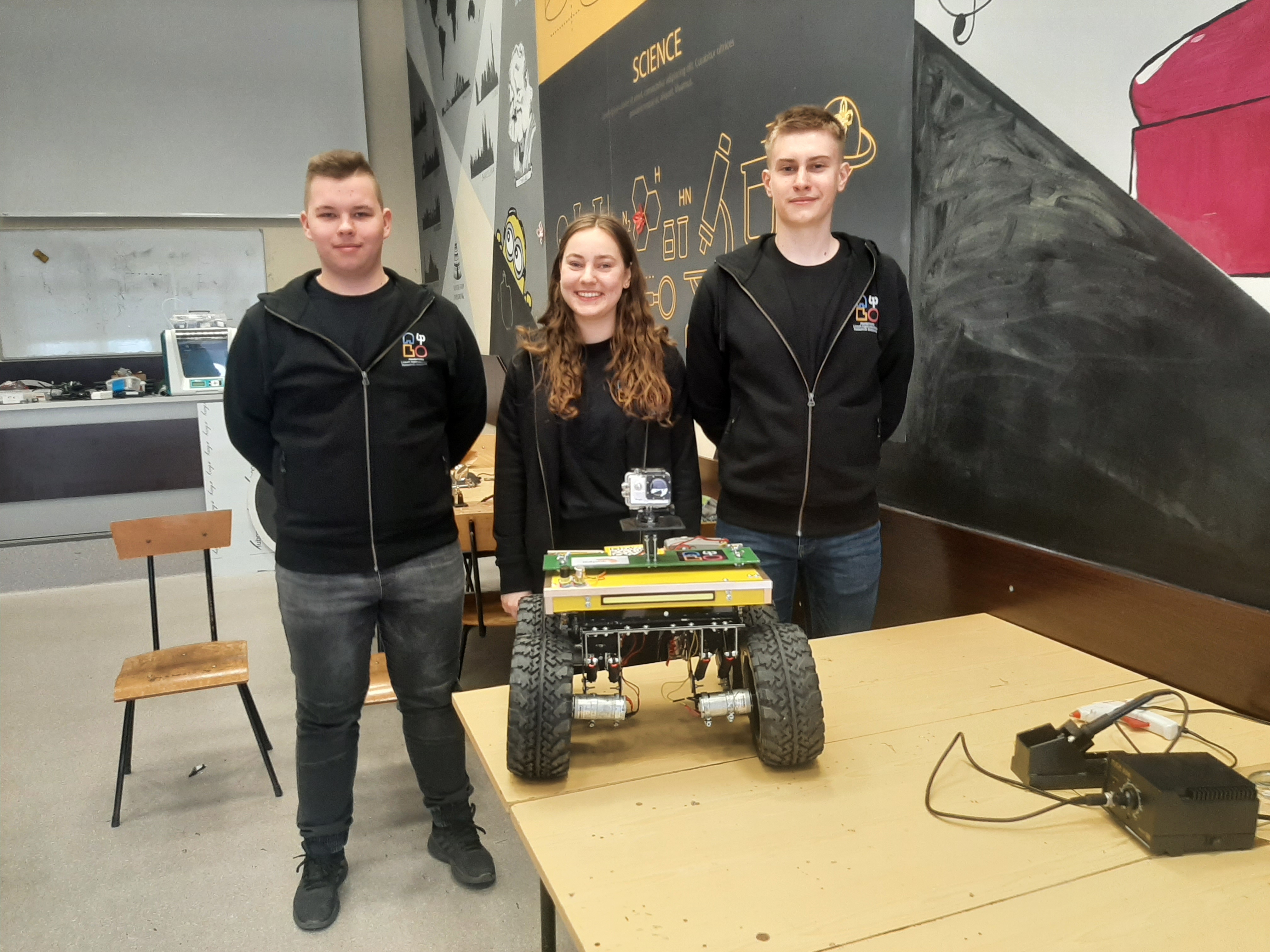 Trzy młode osoby, po bokach chłopaki, w środku dziewczyna stoją w pracowni. Przed nimi na stole stoi robot, które samodzielnie stworzyli