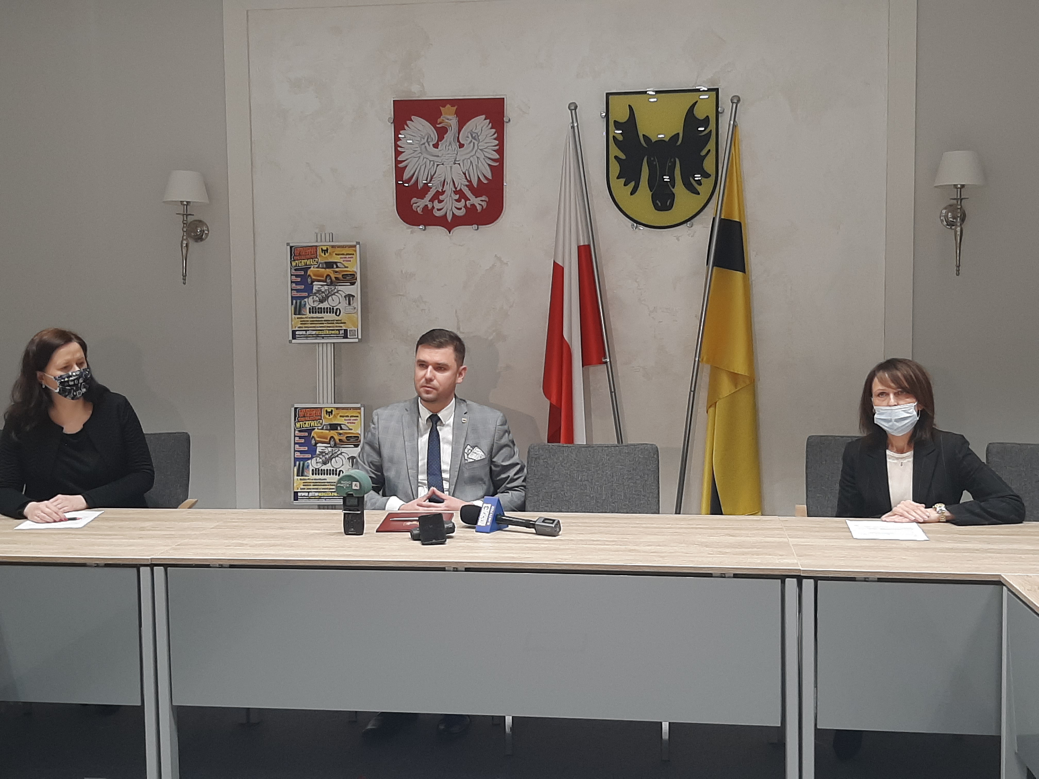 Przy dwóch końcach stołu siedzą kobiety, pośrodku mężczyzna. Na ścianie wisi godło Polski i Wasilkowa