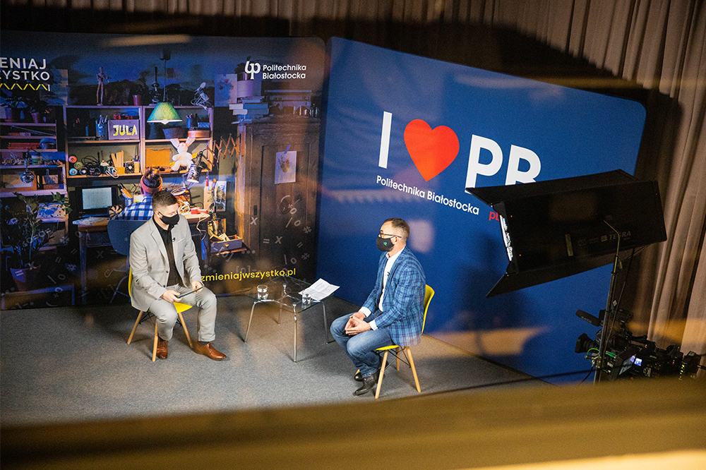 Studio telewizyjne, z góry widoczni są dwaj mężczyźni, którzy ze sobą rozmawiają. Po prawej stronie jest niebieska ścianka Politechniki Białostockiej z hasłem: "I love PB"