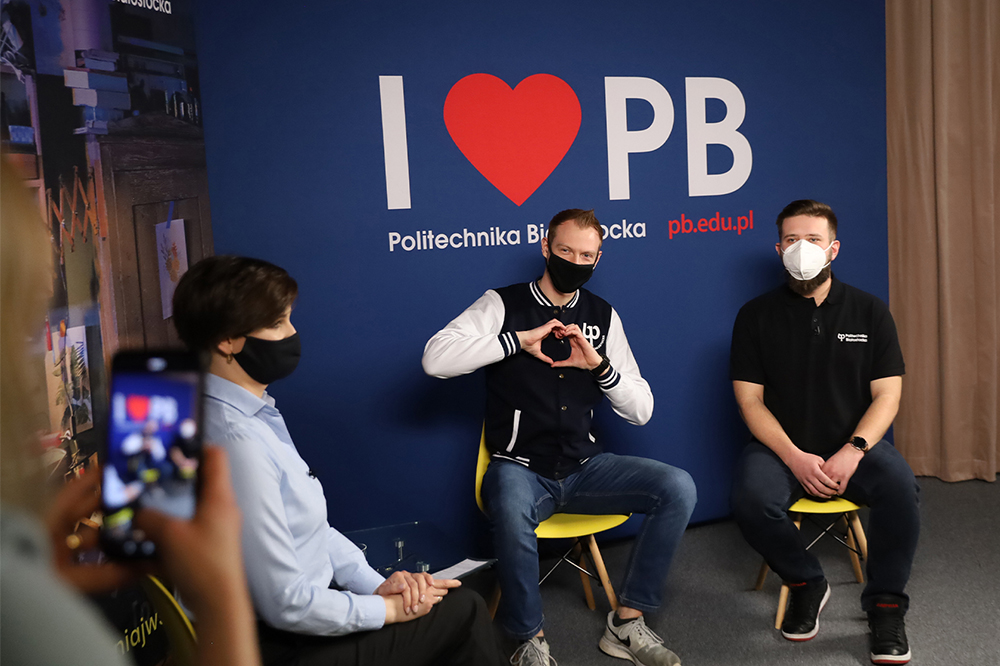 Dwóch studentów siedzi na krzesłach na tle ścianki, na której jest napis: "I love PB". Chłopak siedzący po lewej strony układa dłonie w kształt serca, również z lewej strony siedzi kobieta, która ma na sobie jasną koszulę oraz na krótko ścięte włosy. Tym trzem osobom, dziewczyna robi zdjęcia telefonem. Kobieta znajduje się w lewym rogu