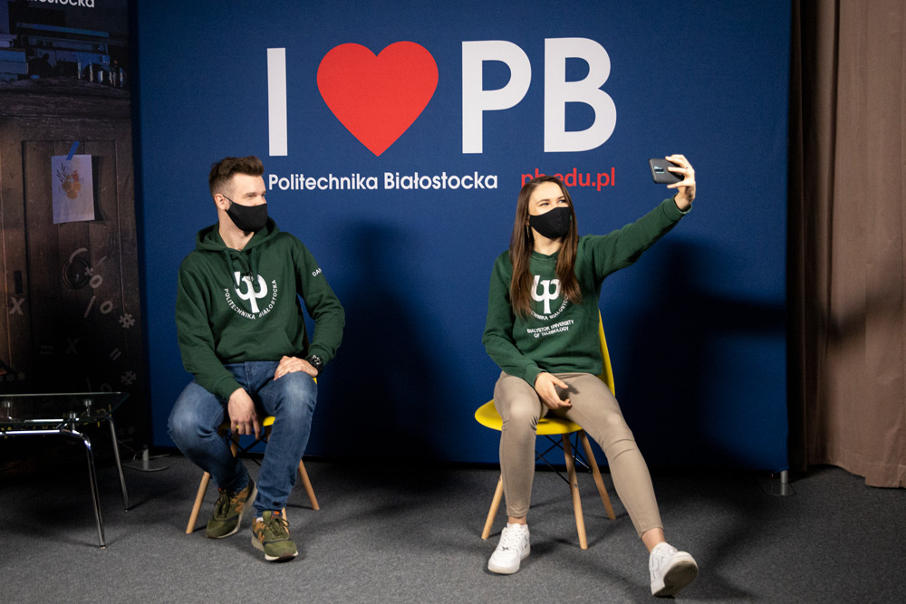 Student i studentka siedzą na krzesłach na tle niebieskiej ścianki z napisem "I love PB". Ubrani są w zielone bluzy z białym logiem PB. Dziewczyna, która znajduje się po prawej stronie robi selfie telefonem komórkowym