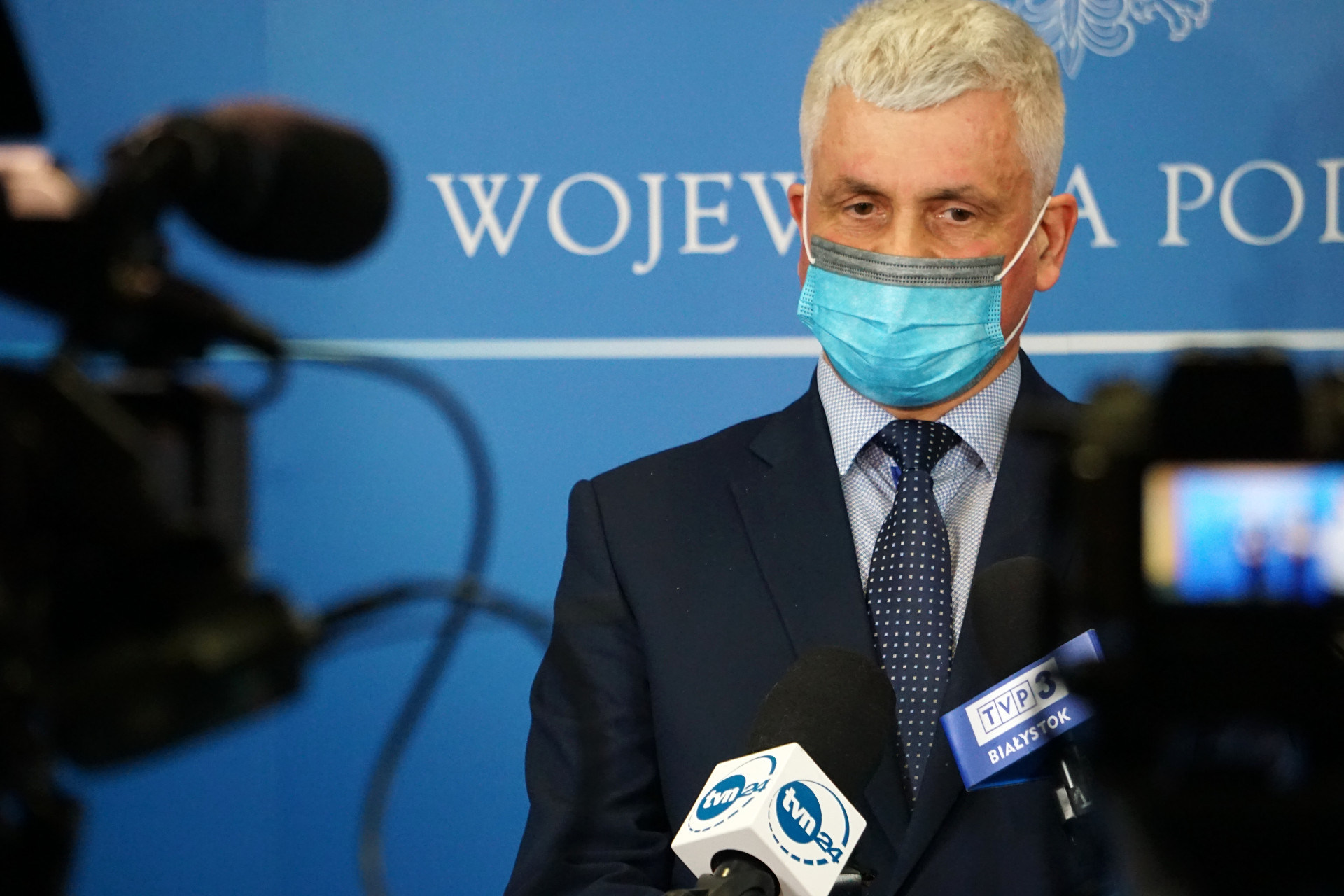 Wojewoda podlaski jest w ciemnym garniturze stoi na tle niebieskiej ścianki. Kadr pochodzi z konferencji prasowej, przed wojewoda są mikrofony oraz kamery