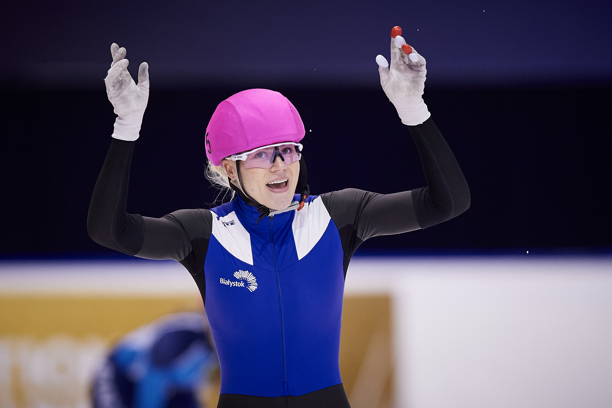 Zadowolona zawodniczka short tracka Natalia Maliszewska. Kobieta ma różowy kask, okularki i niebiesko - czarno z białymi dodatkami kombinezon