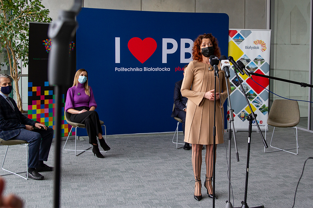 Kobieta w beżowej sukience stoi przed mikrofonami, za nią widoczna jest granatowa ścianka z napisem I love PB. Po lewej stronie w fioletowej bluzce siedzi kobieta
