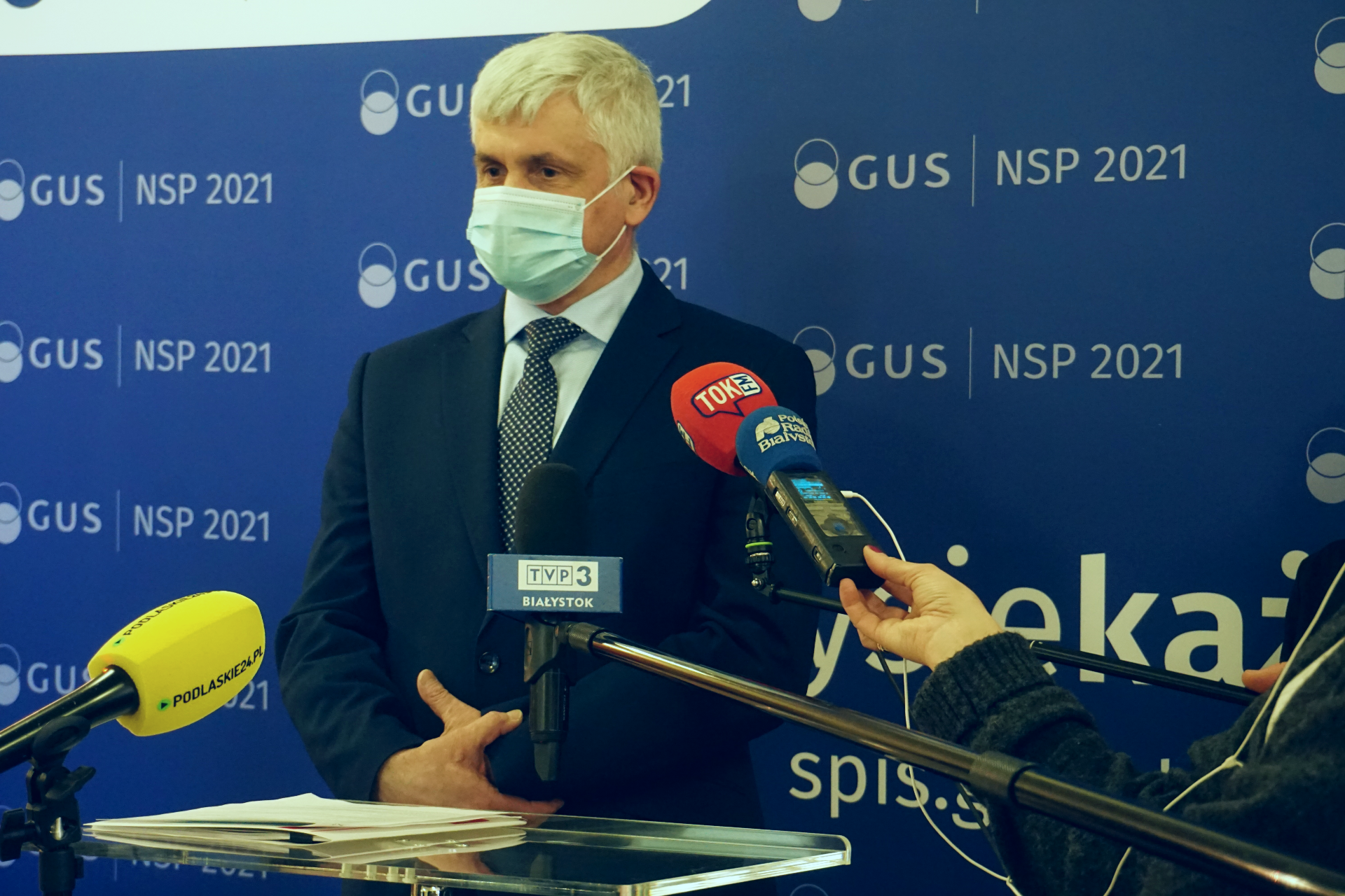 Mężczyzna w garniturze stoi na tle niebieskiej ścianki. Przed nimi dziennikarze trzymają mikrofony