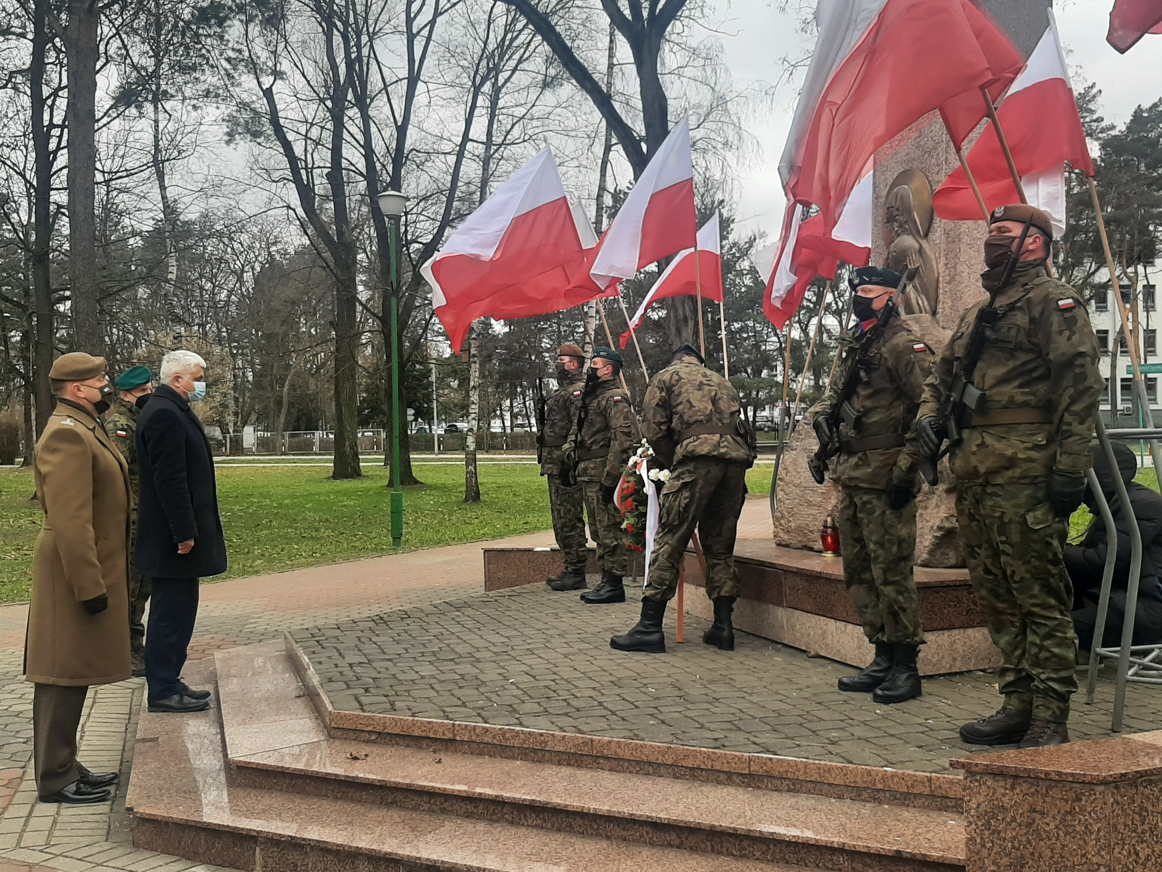 Przy pomniku powiewają biało- czerwone flagi Polski. Przy pomniku żołnierze trzymają wartę. Po lewej stronie wojewoda i wojskowy stoją przed Pomnikiem Katyńskim