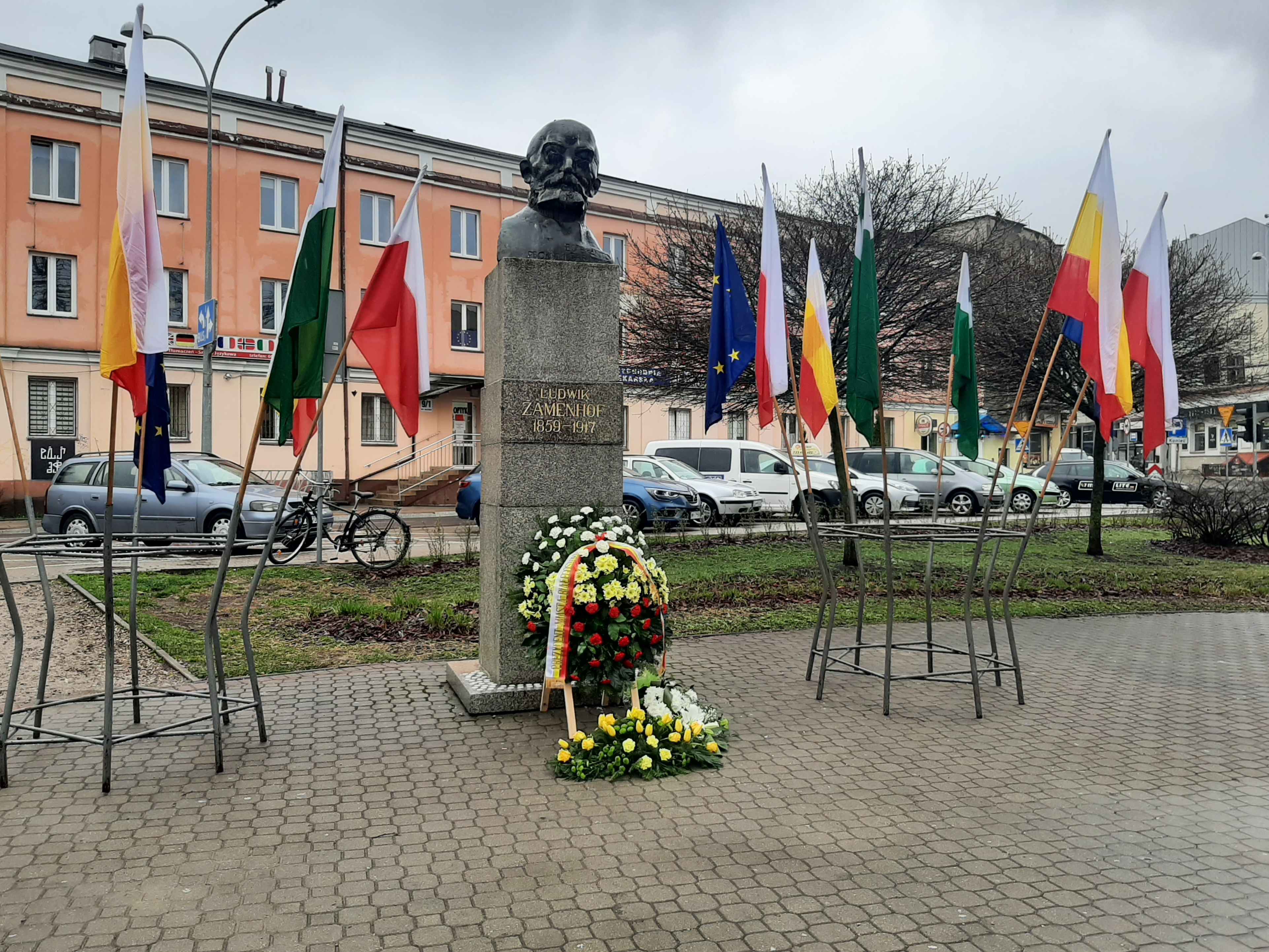 Pomnik, głowa na postumencie, Ludwika Zamenhofaprzy ul. Malmeda. Wokół pomnika znajdują się stojaki, w nich flagi: Polski, Białegostoku, Esperantystów. Przed pomnikiem leżą kwiaty