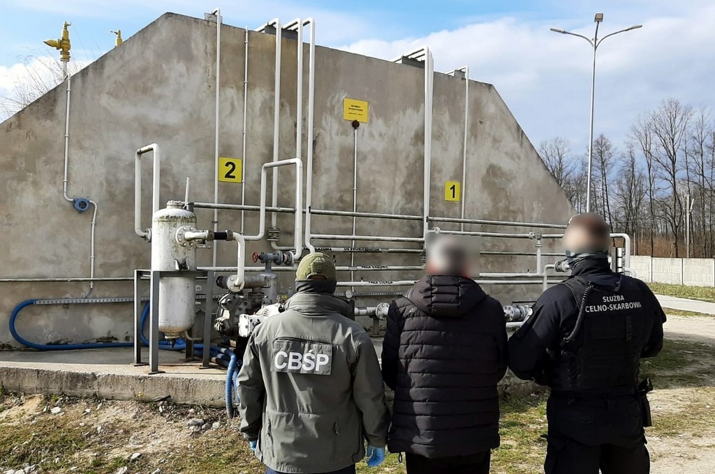 Trzy osoby przed aparaturą przesyłową gazu, z lewej stoi funkcjonariusz CBŚP z prawej funkcjonariusz Służby Celno-Skarbowej