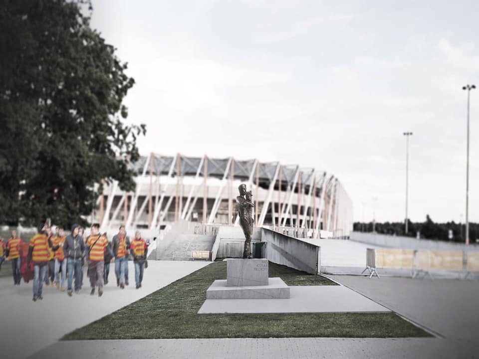 W oddali widać Stadion Miejski w Białymstoku.. Z lewej strony idą kibice w koszulkach w żółto - czerwone paski. Na środku zdjęcia znajduje się pomnik 42. Pułku Piechoty