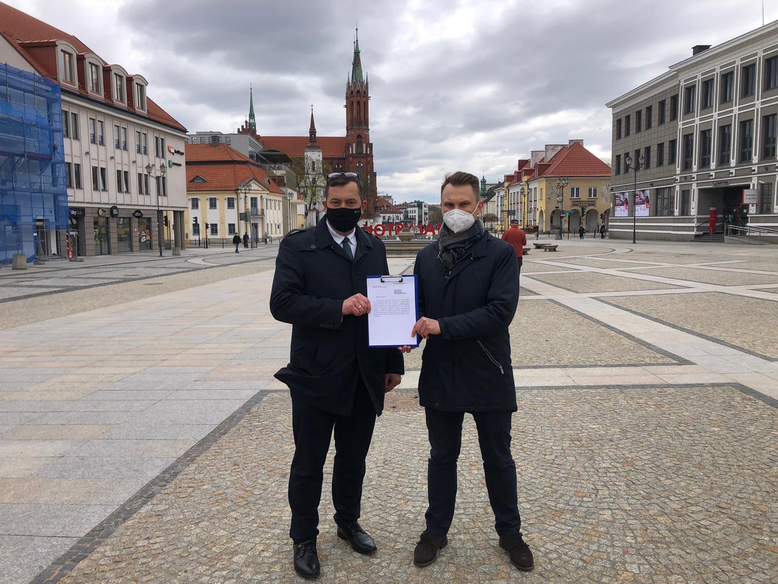 Dwóch posłów; po lewej stronie - Stefan Krajewski, z prawej Krzysztof Truskolaski trzymają wspólnie wniosek, który przygotowali. Mężczyźni stoją na Rynku Kościuszki w Białymstoku, za nimi widać Katedrę