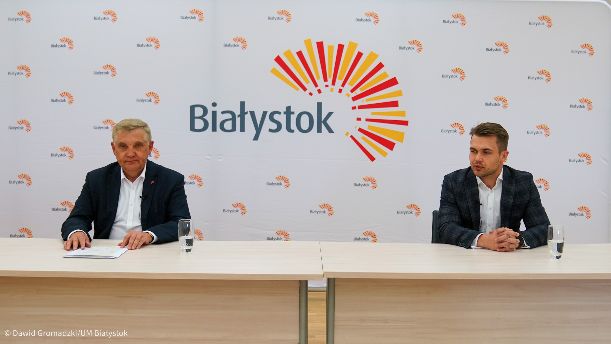 Dwóch mężczyzn siedzi przy stole na tle ścianki z napisem Białystok i z logiem wschodzącego słońca. Z lewej strony siedzi prezydent Białegostoku.