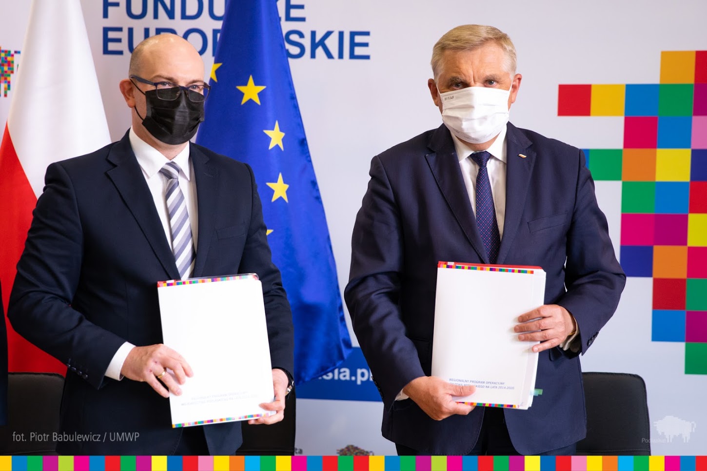 Z lewej strony stoi marszałek Artur Kosicki, z prawej prezydent Białegostoku. Obaj mężczyźni mają ciemne garnitury i maseczki ochronne na twarzy. Za nimi stoi flaga Unii Europejskiej. Z prawej strony widać fragment loga województwa podlaskiego - pikselowego żubra
