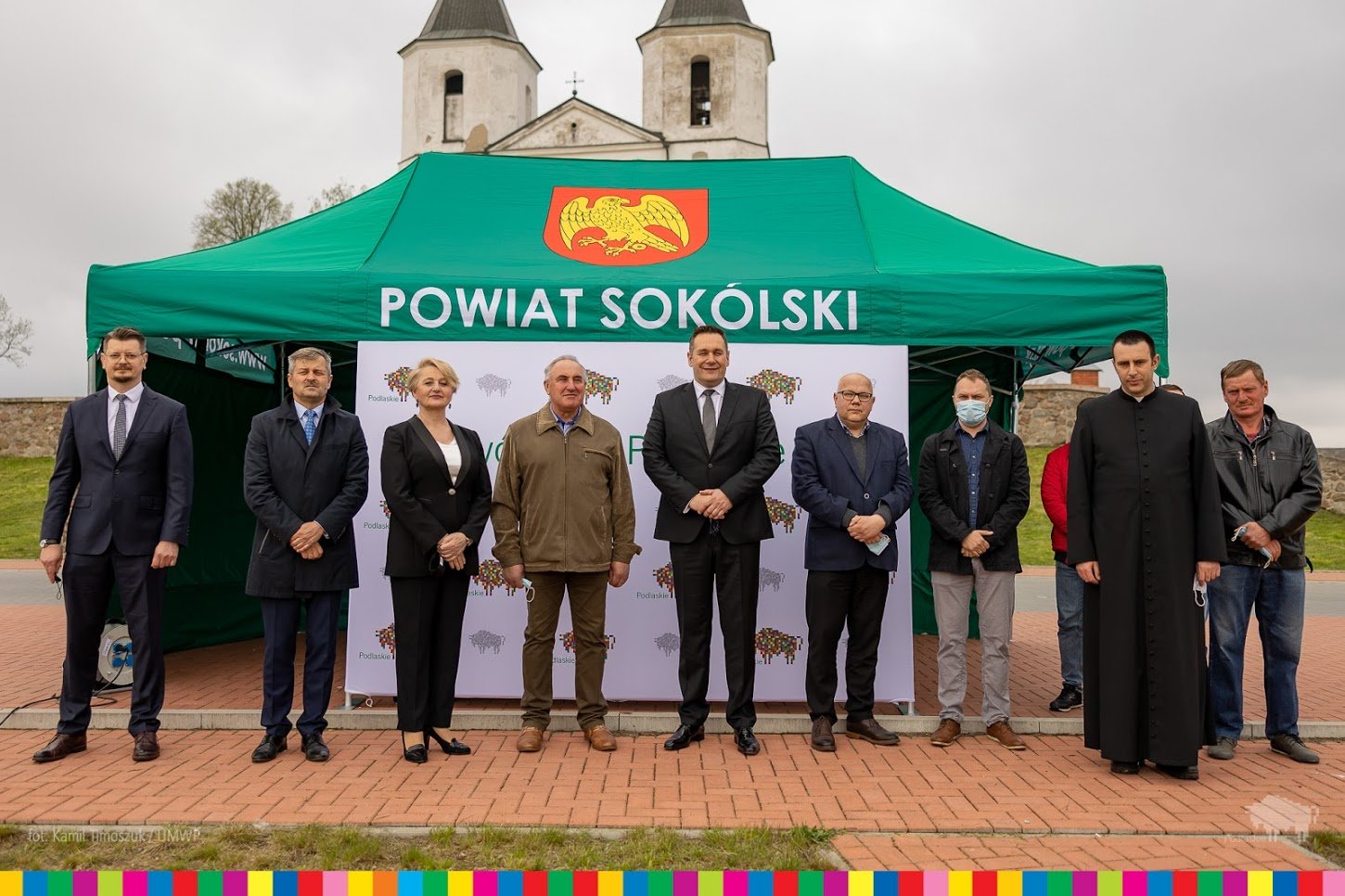 Ludzie stoją w rzędzie za nimi stoi zielony namiot z napisem Powiat Sokólski oraz kościół