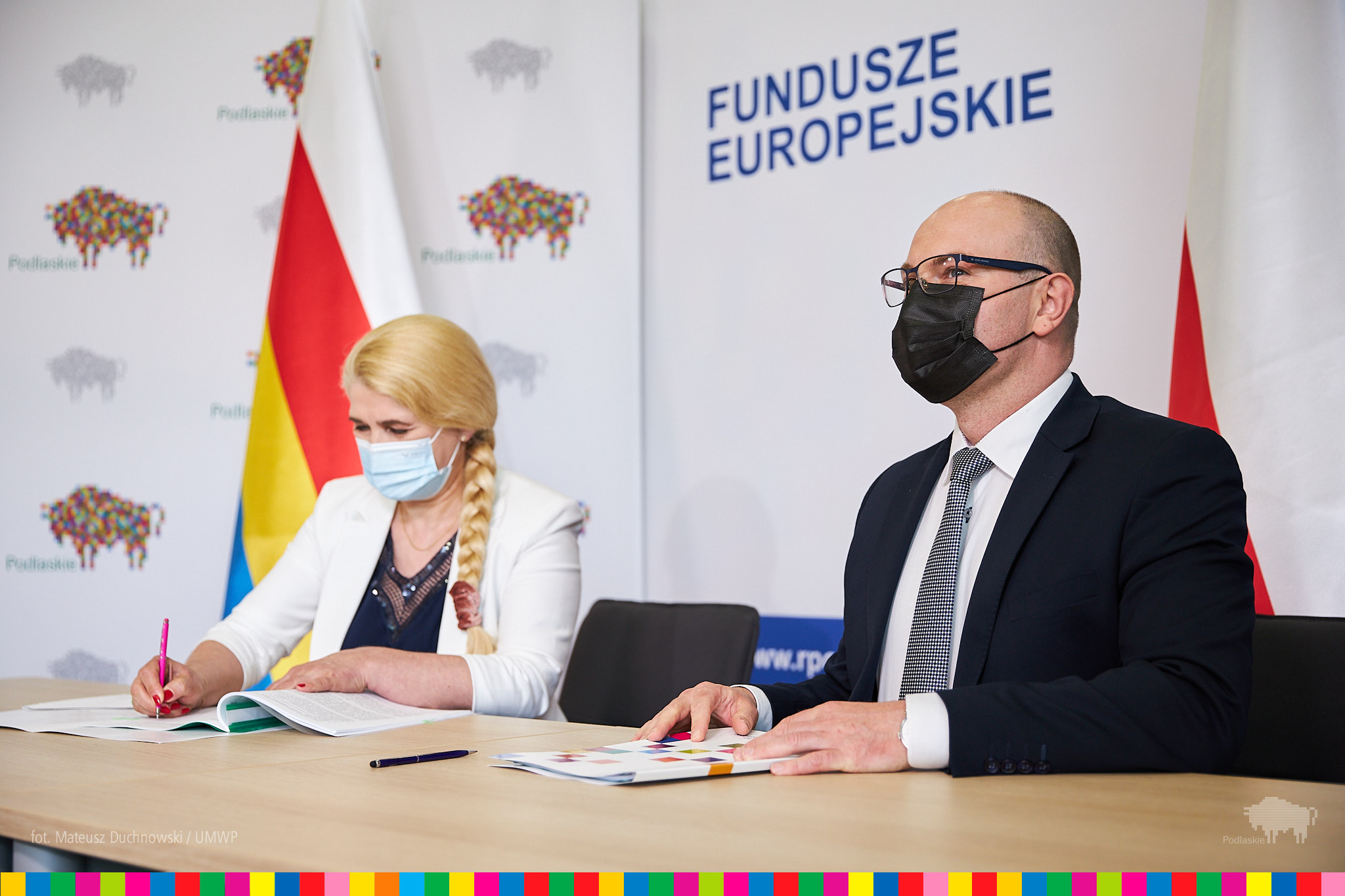 Przy stole, od lewej strony siedzi kobieta i podpisuje umowę, z prawej mężczyzna, który patrzy przed siebie. Za nimi na ścianie jest napisane "Fundusze Europejskie", ścianka województwa w żubry i flaga województwa: biało - czerwono - żółto - niebieska