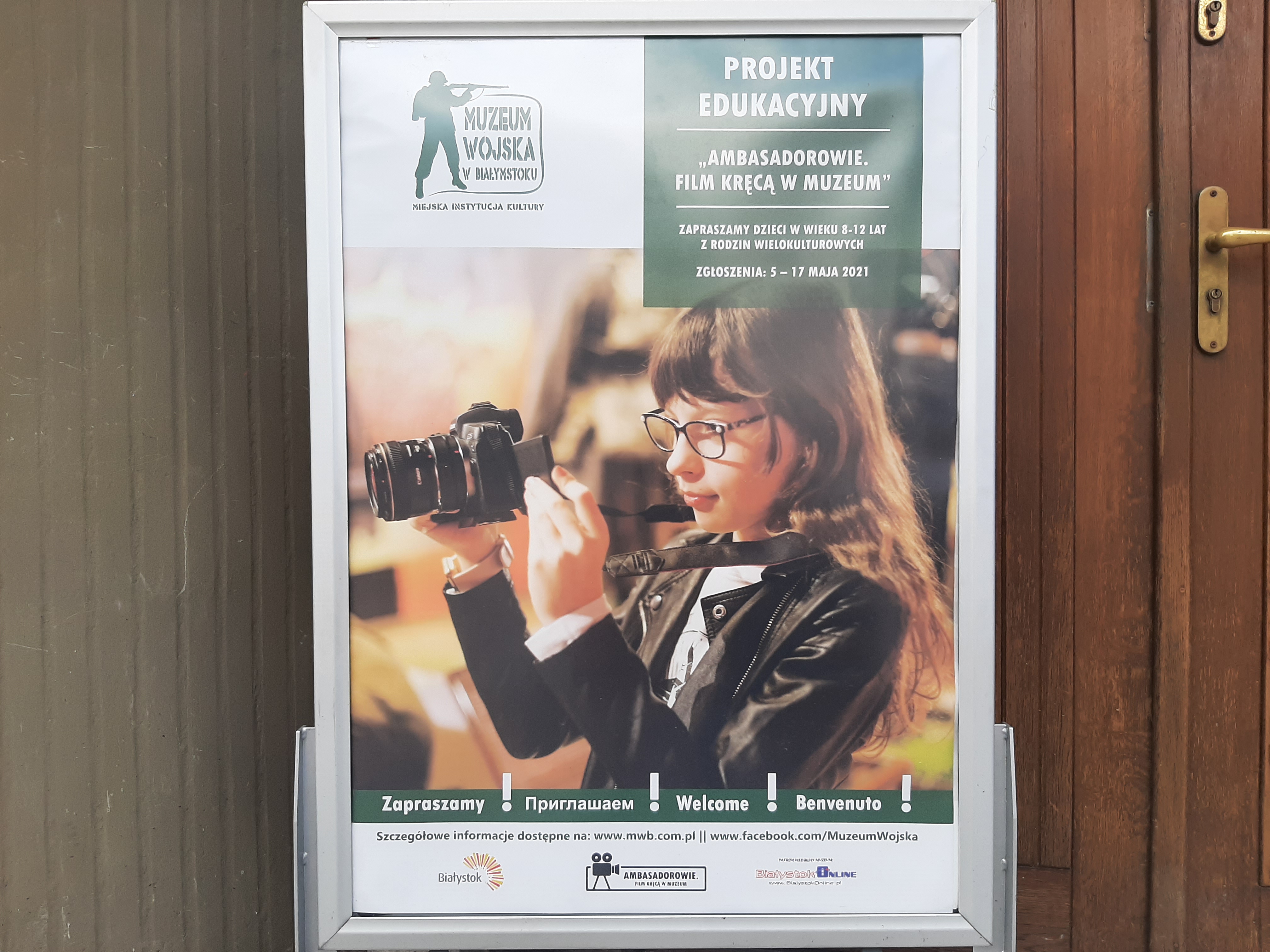 Przy drzwiach stoi plakat, na którym widoczna jest dziewczynka z aparatem na szyi i która patrzy w ekran urządzenia