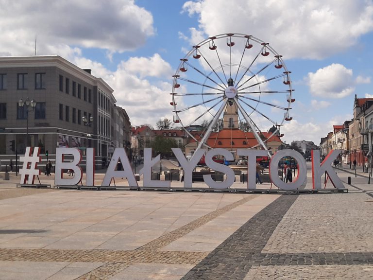 Okolice Rynku Kościuszki w Białymstoku, centralnie na placu ustawiony jest biały napis hasztag Białystok, za napisem widoczna jest karuzela typu diabelski młyn