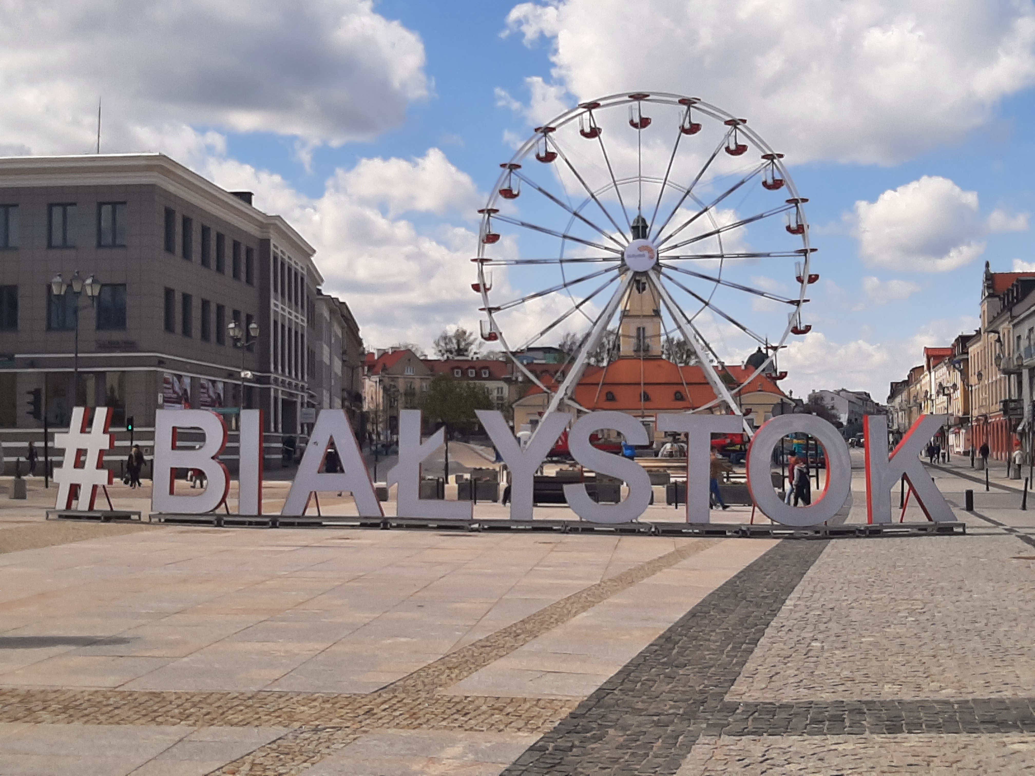 Okolice Rynku Kościuszki w Białymstoku, centralnie na placu ustawiony jest biały napis hasztag Białystok, za napisem widoczna jest karuzela typu diabelski młyn