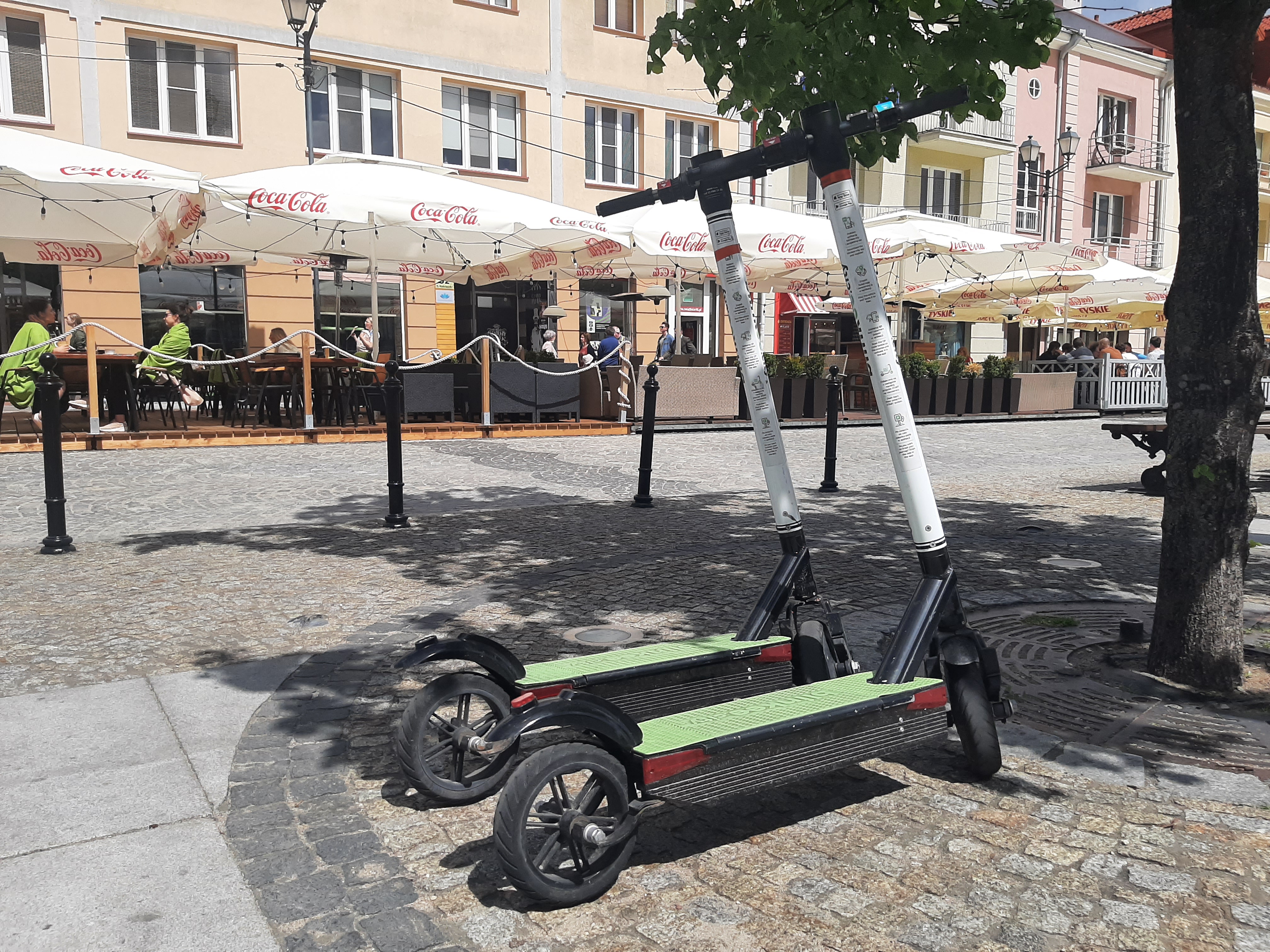 Pod drzewem stoją dwie hulajnogi elektryczne. W oddali znajdują się ogródki gastronomiczne, które są ustawione w centrum Białegostoku