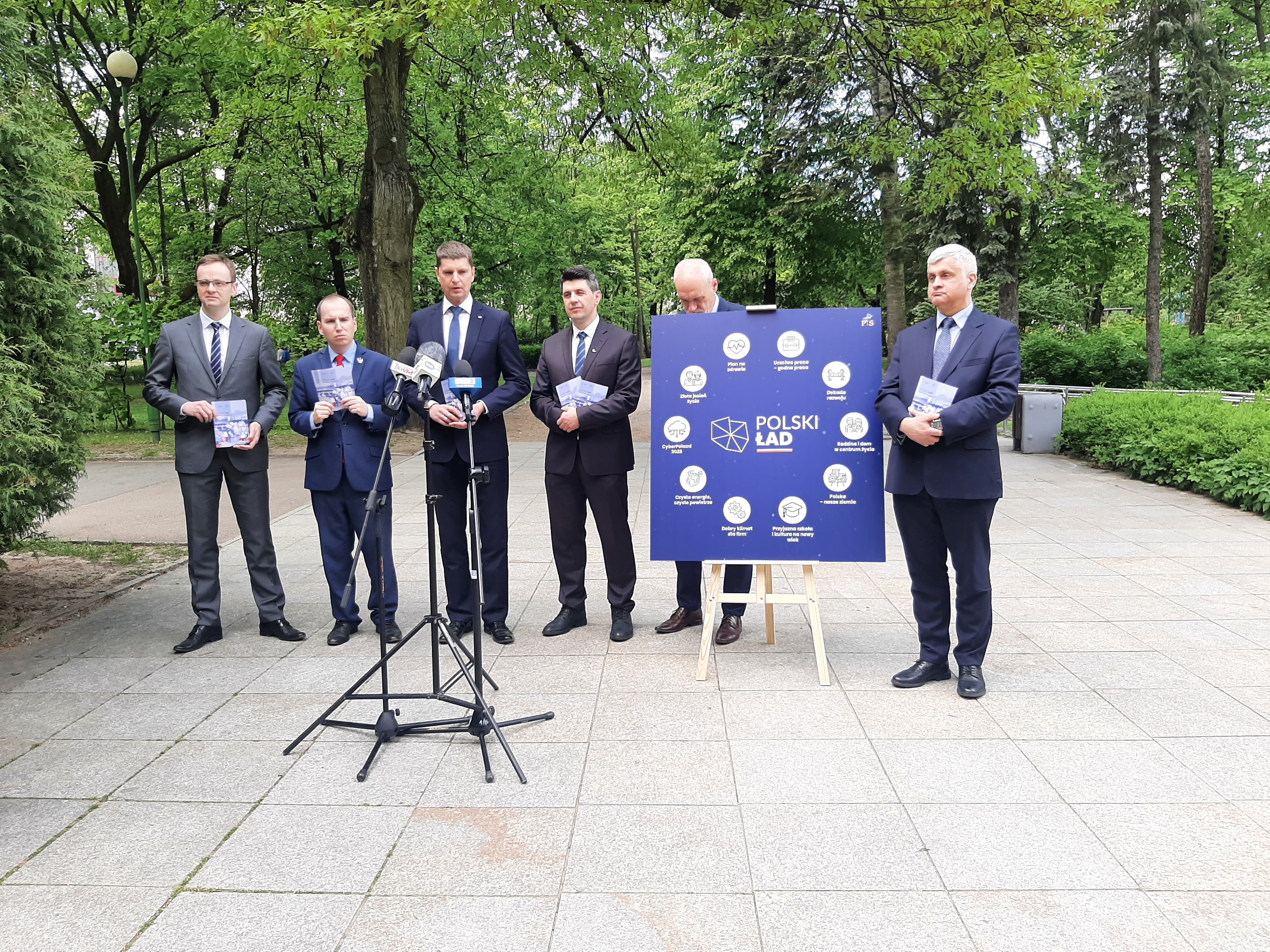 W Parku Plany, w rzędzie stoją politycy Prawa i Sprawiedliwości. Między nimi stoi niebieski plakat
