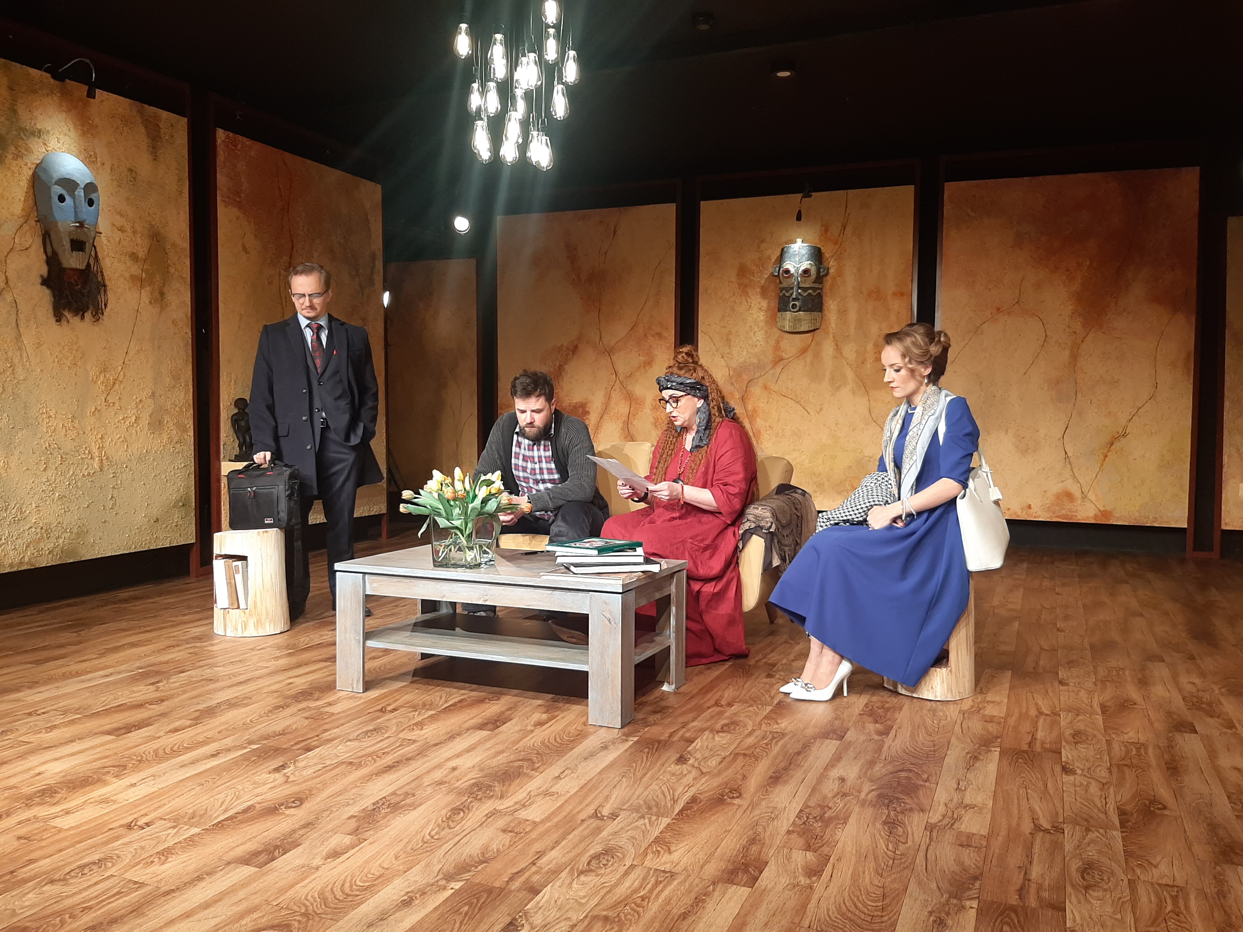 Fragment próby teatralnej. Przy stoliku, na kanapie siedzą dwie osoby. Z lewej stoi mężczyzna w płaszczy i z prawej siedzi kobieta w niebieskiej sukience