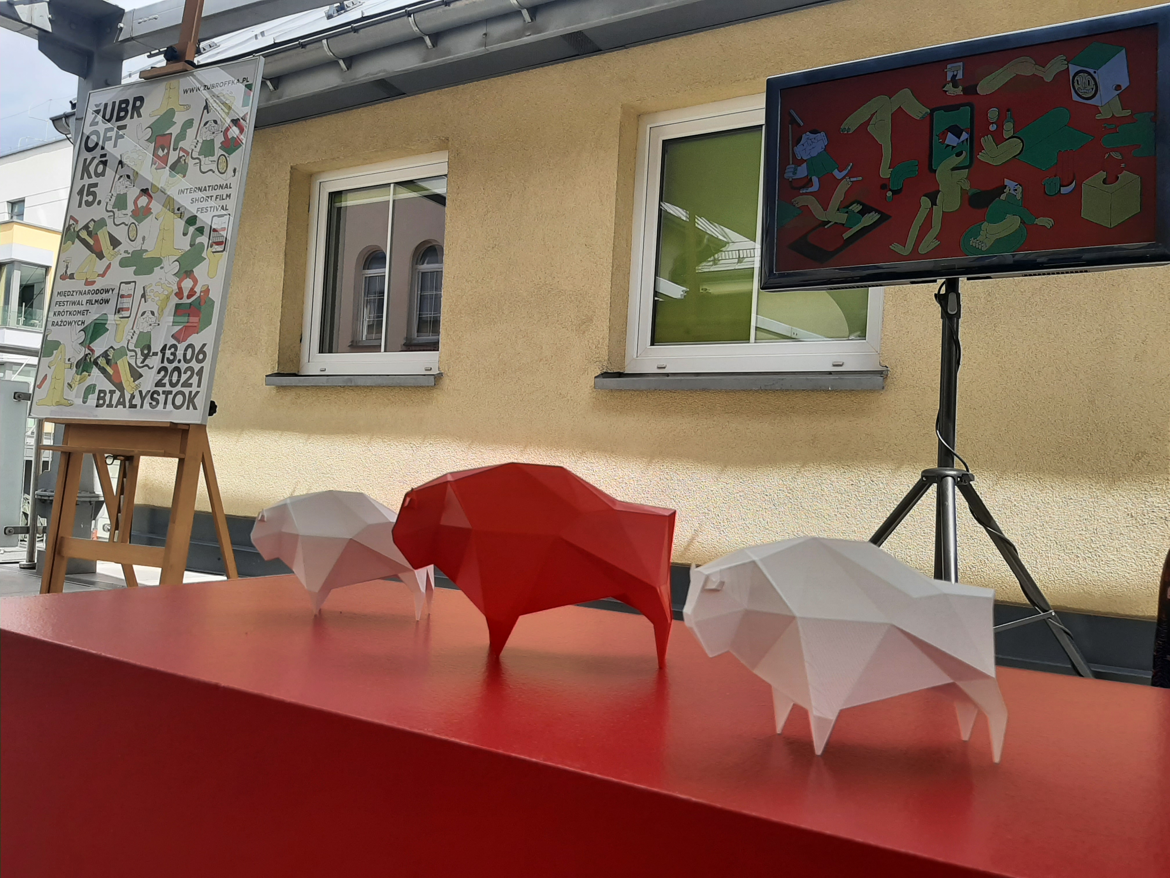 Na czerwonym stole stoją trzy plastikowe żubry; biały, czerwony, biały. W oddali, z lewej strony na sztaludze stoi plakat, z prawej telewizor
