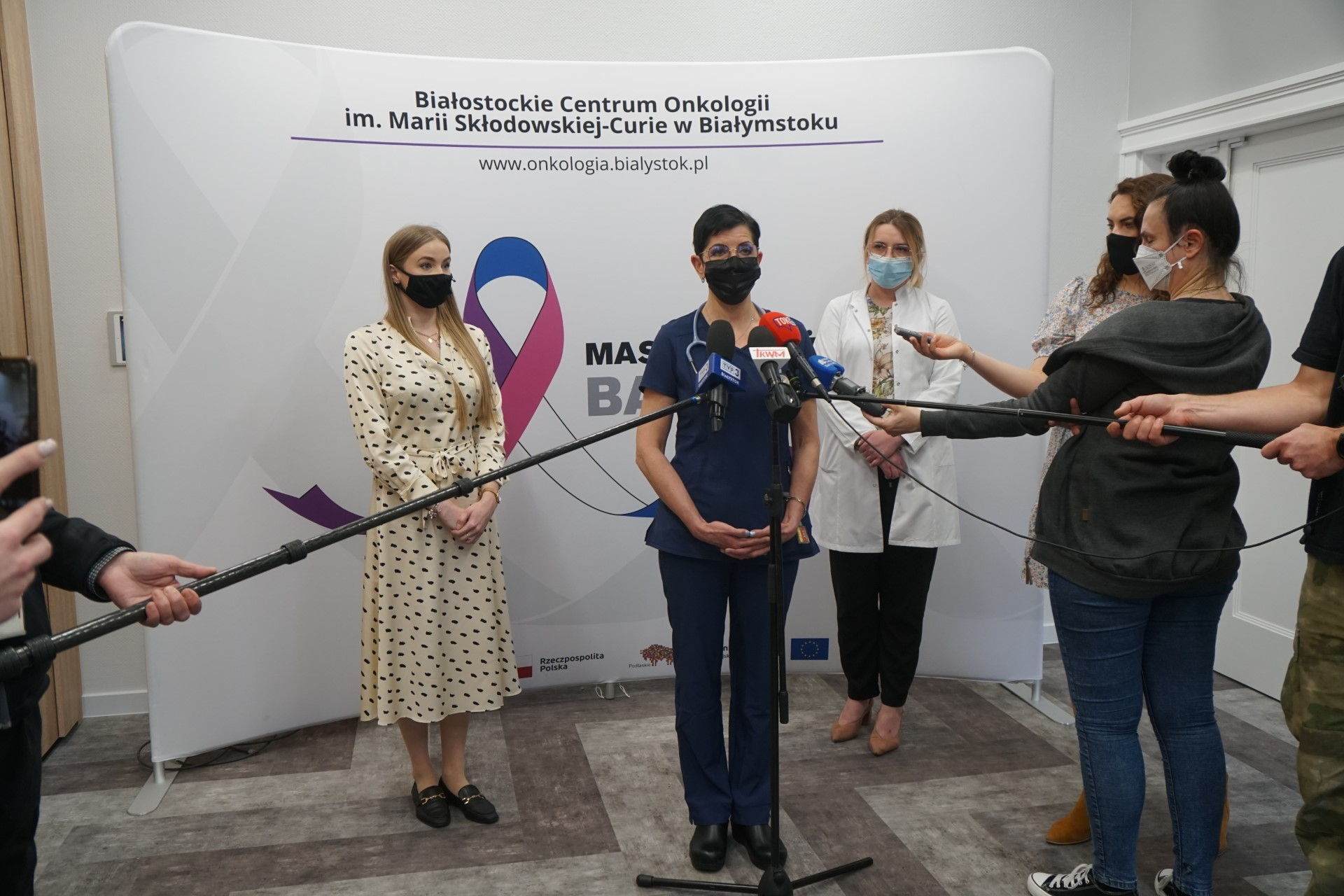 Kobieta mówi do mikrofonów dziennikarzy. Za nią, na ściance Białostockiego Centrum Onkologii, stoją jeszcze dwie kobiety