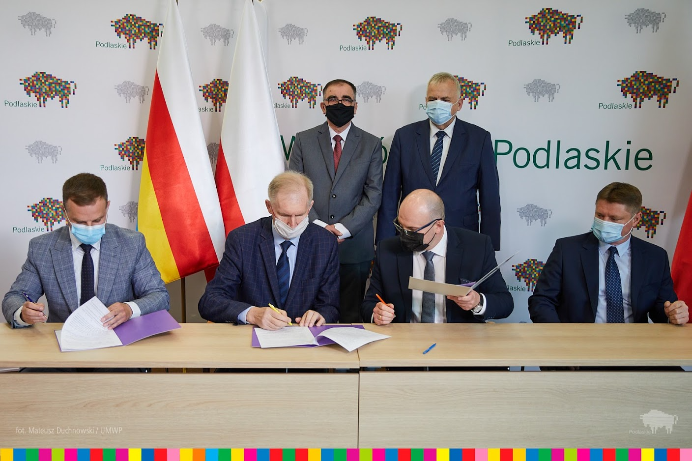 Czterech mężczyzn siedzi przy stole, podpisują umowy. Za nimi stoją jeszcze dwie osoby. W tle widać ściankę z logo województwa. symbolem regionu jest pikselowy żubr