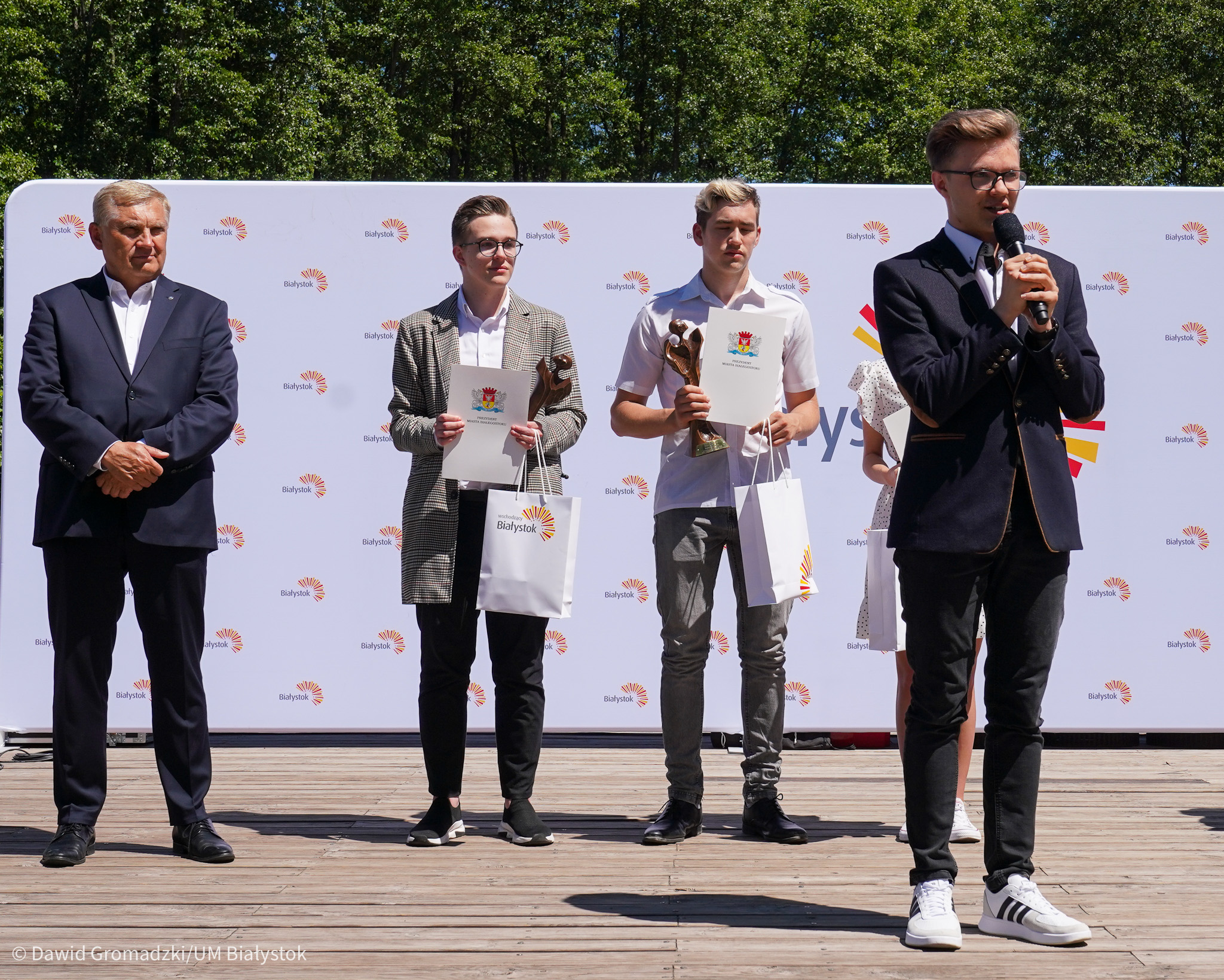 Na scenie, na tle ścianki z logo Białegostoku, wschodzącym słońcem stoją, od lewej prezydent miasta. Obok niego stoją laureaci konkursu. Przed nimi stoi chłopak, który mówi przez mikrofon