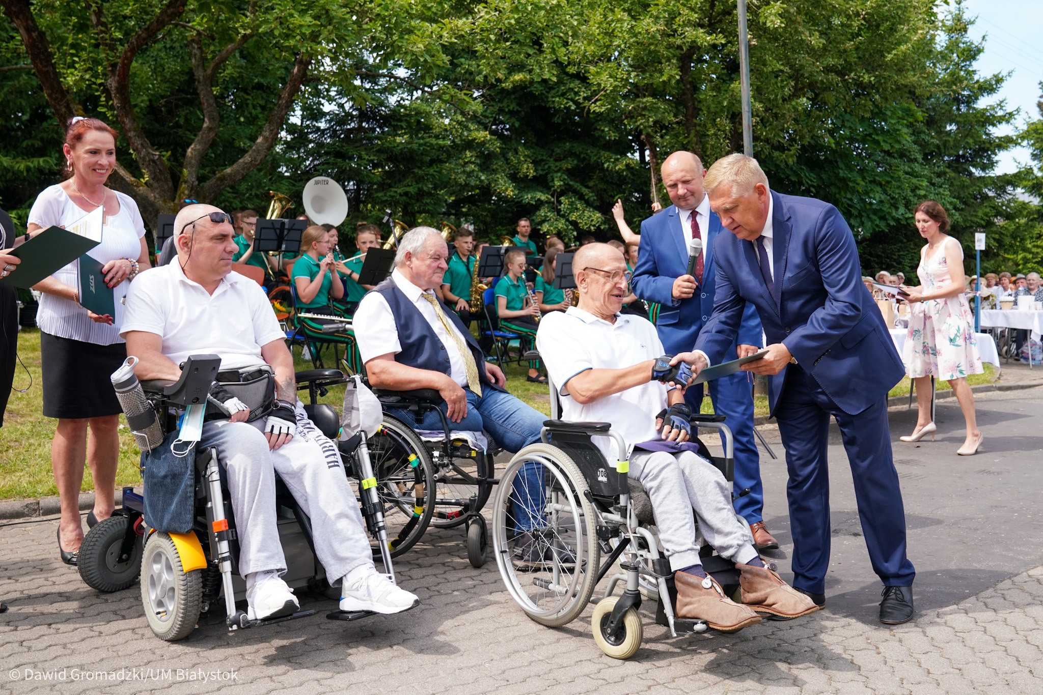 Trzech mężczyzn na wózkach, jednemu z nich prezydent Białegostoku ściska dłoń i wręcza nominację. Obok prezydenta stoi dyrektor DPS -u. W tle widoczna jest orkiestra