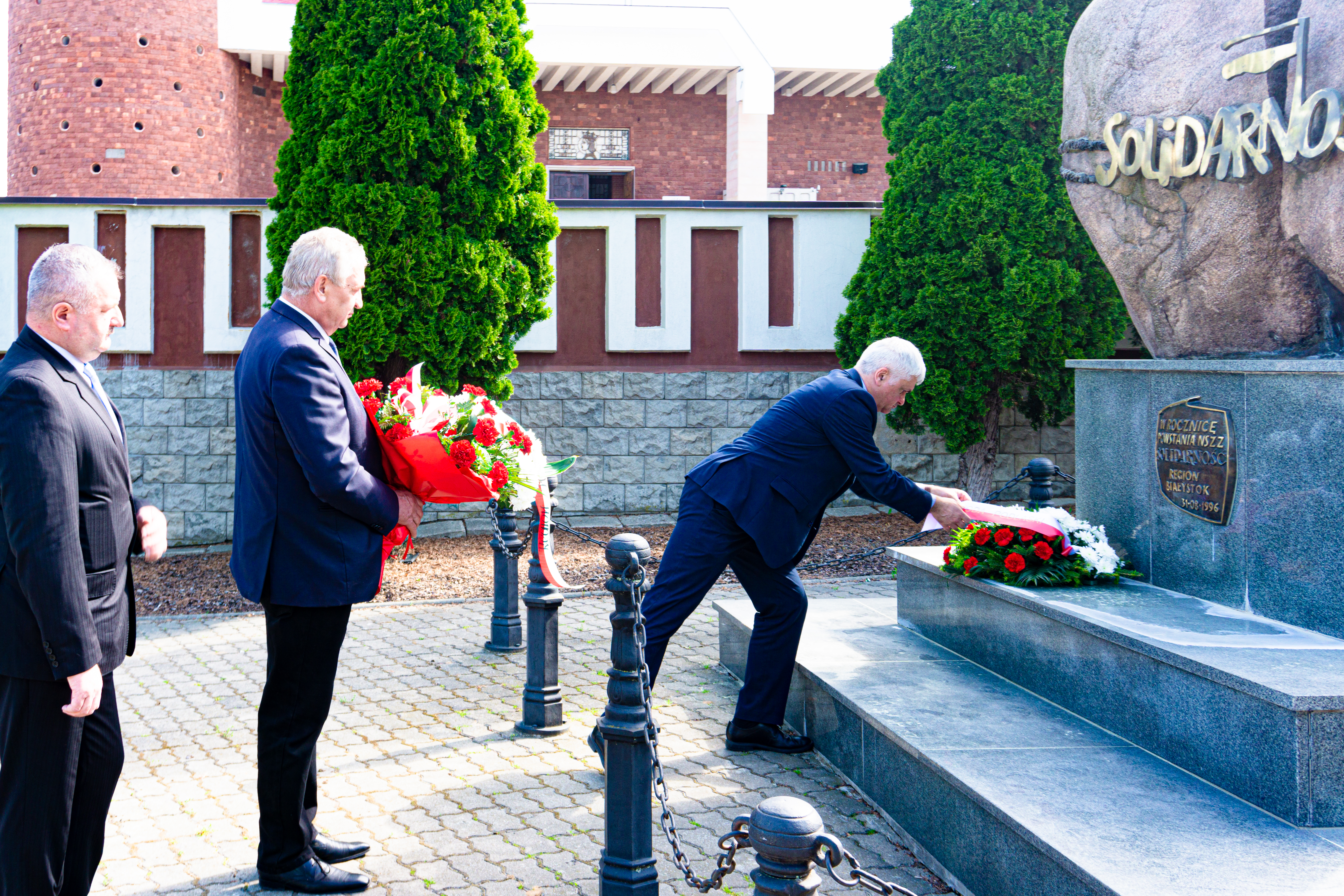 Trzech mężczyzn znajduje się przed pomnikiem Solidarności w Białymstoku. Wojewoda, który stoi jako pierwszy poprawia wstęgę na złożonym wieńcu