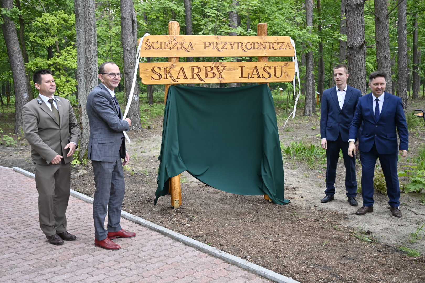 Czterech mężczyzn stoi w lesie, jeden z nich odsłonił drewnianą tablicę z napisem : Ścieżka przyrodnicza. Skarby lasu