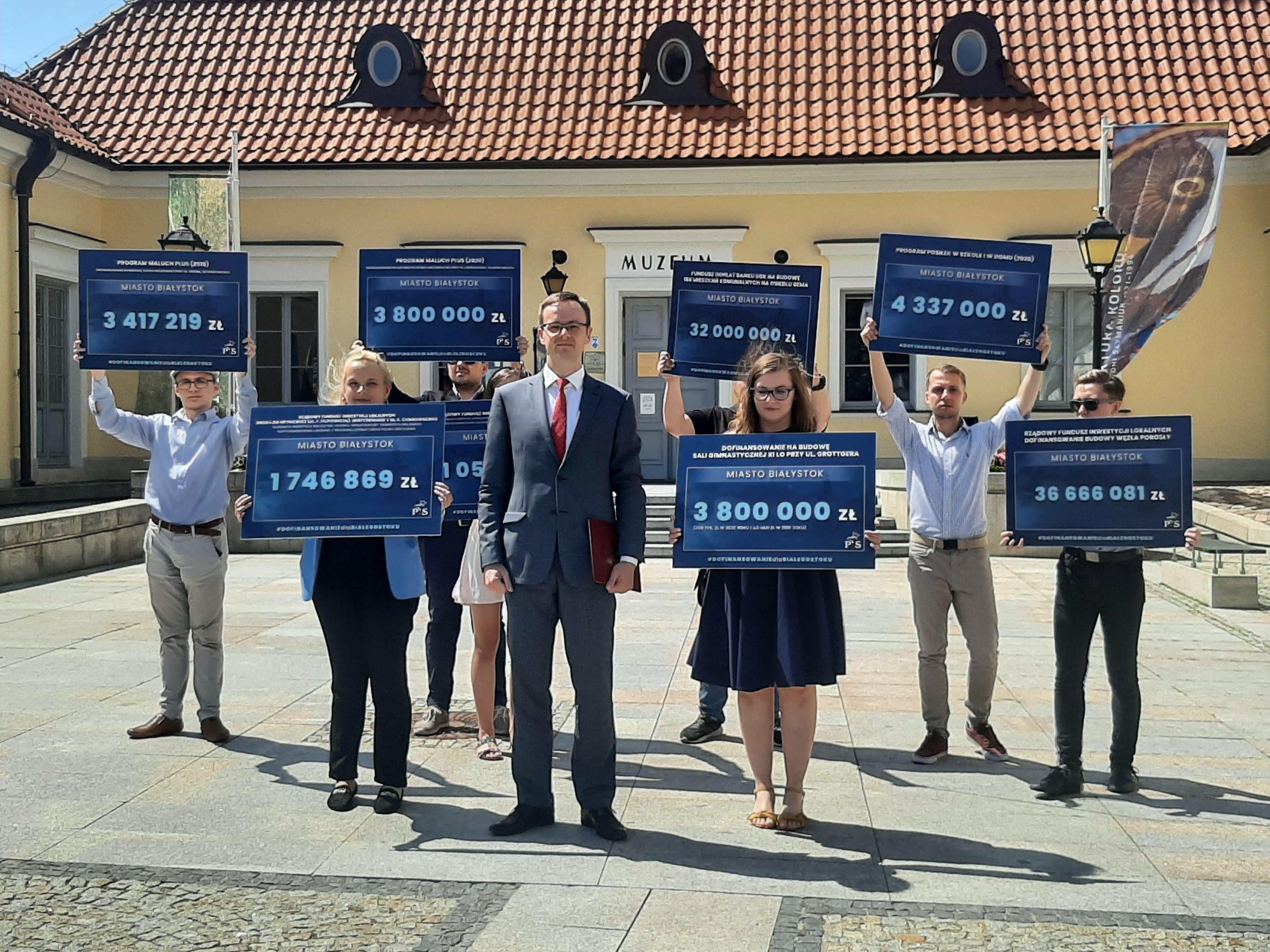 Grupa ludzi stoi na tle Ratusza, w centrum Białegostoku. Osoby te trzymają tabliczki z kwotami dofinansowań, które otrzymał Białystok od rządu