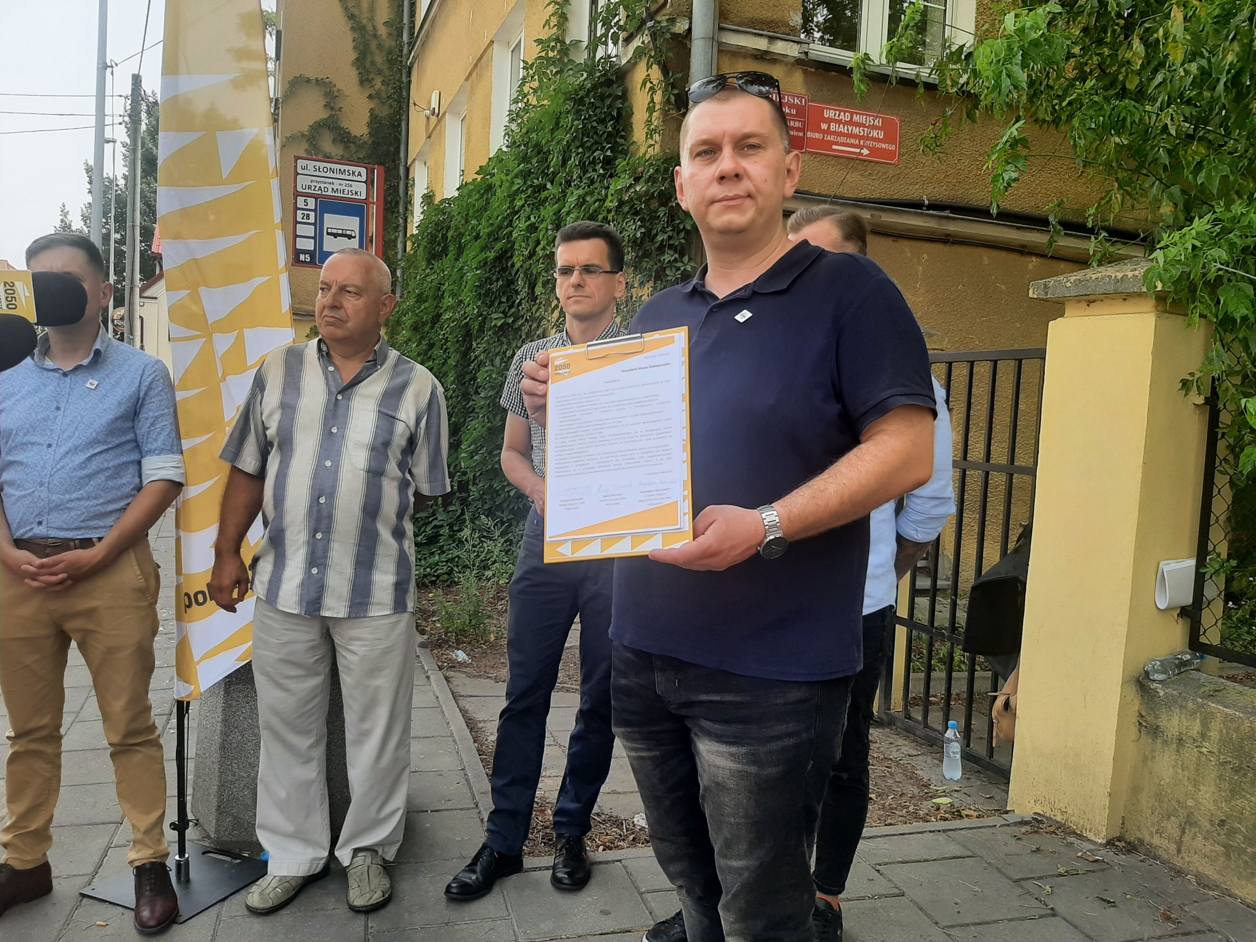 Radny stoi, trzyma w ręku interpelację. Za nim stoją inne osoby