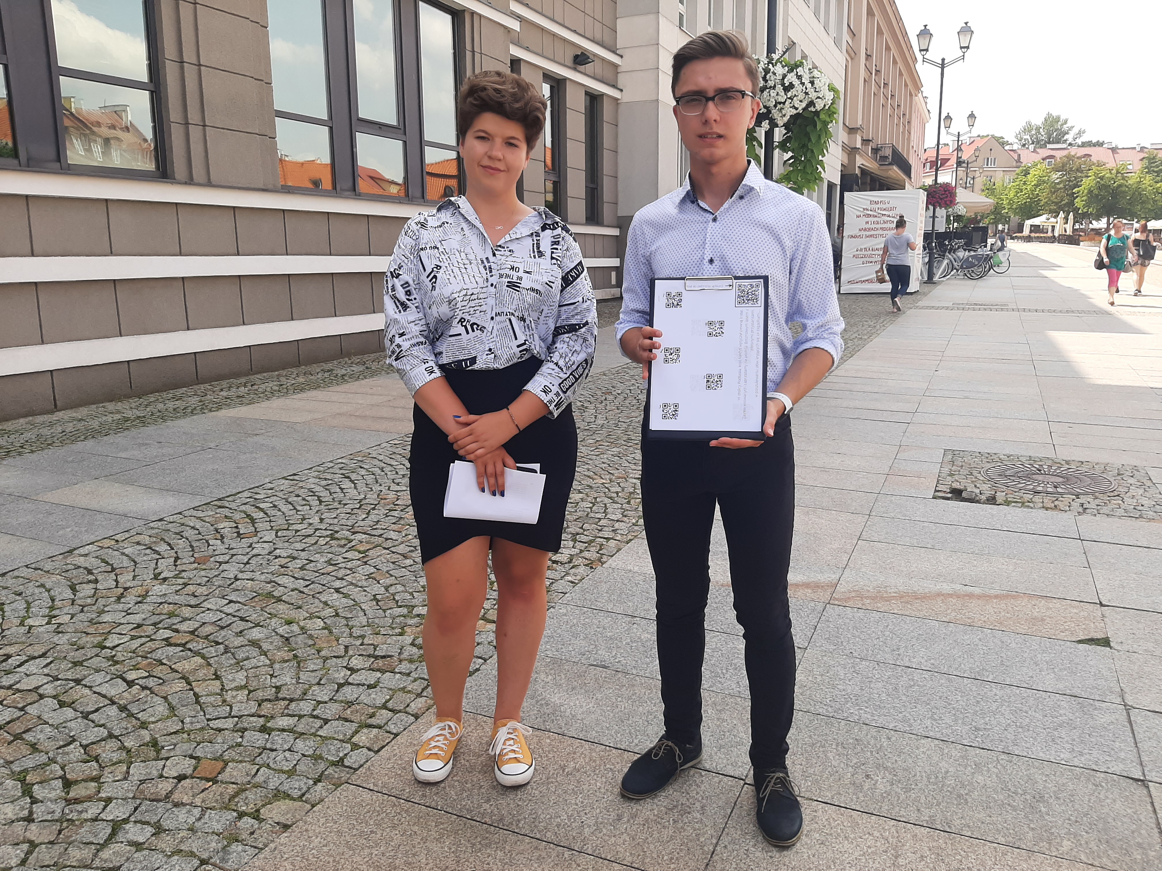 Z lewej strony stoi dziewczyna, z prawej chłopak, który trzyma w ręku kartkę z kodami QR