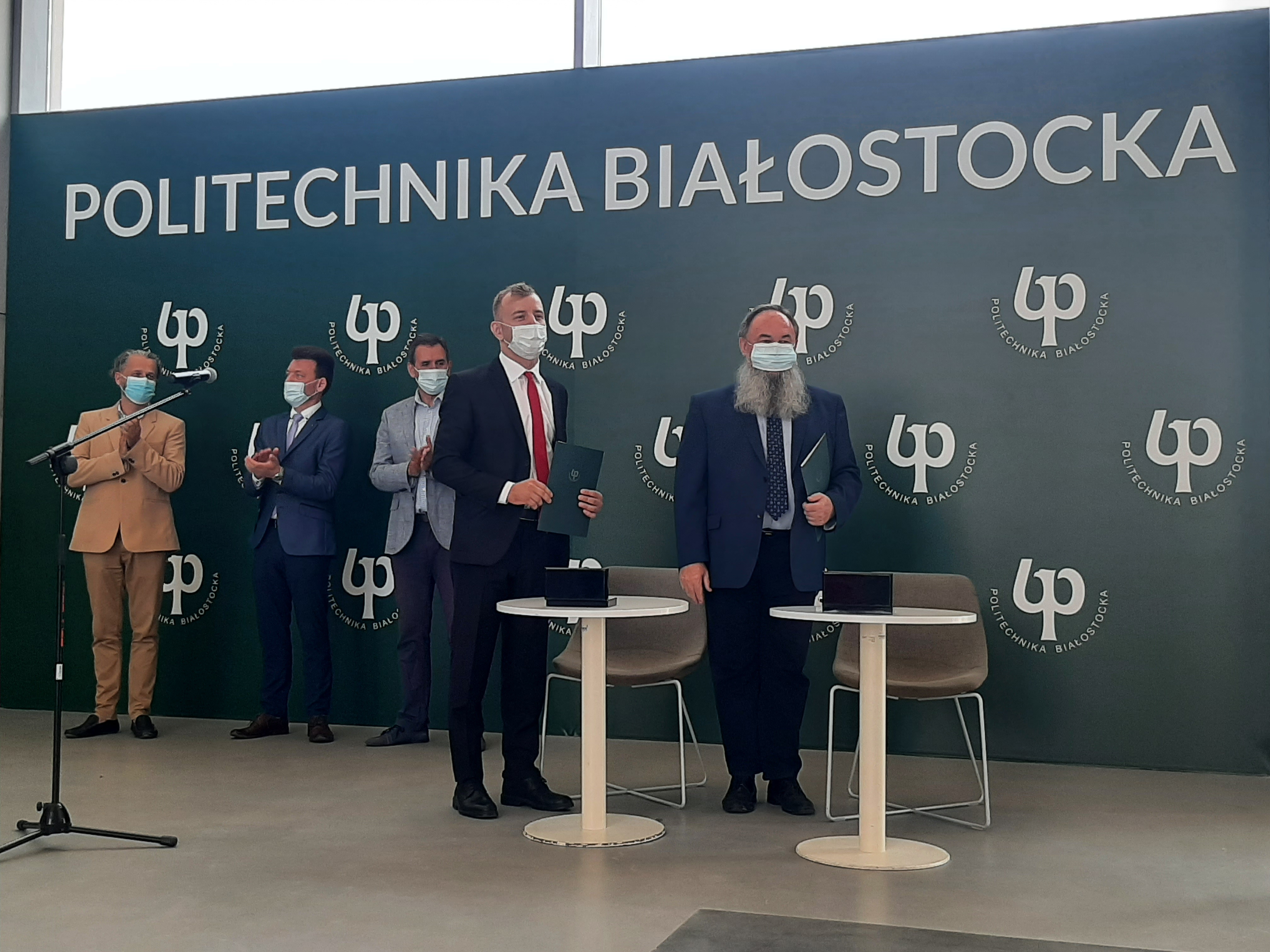 Przy dwóch białych, okrągłych stolikach stoi dwóch mężczyzn. Za nimi widoczna jest zielona ścianka z białym napisem: Politechnika Białostocka i symbolem PB. W poddali widoczni są równej trzej mężczyźni. Wszyscy mają zakryte usta maseczką