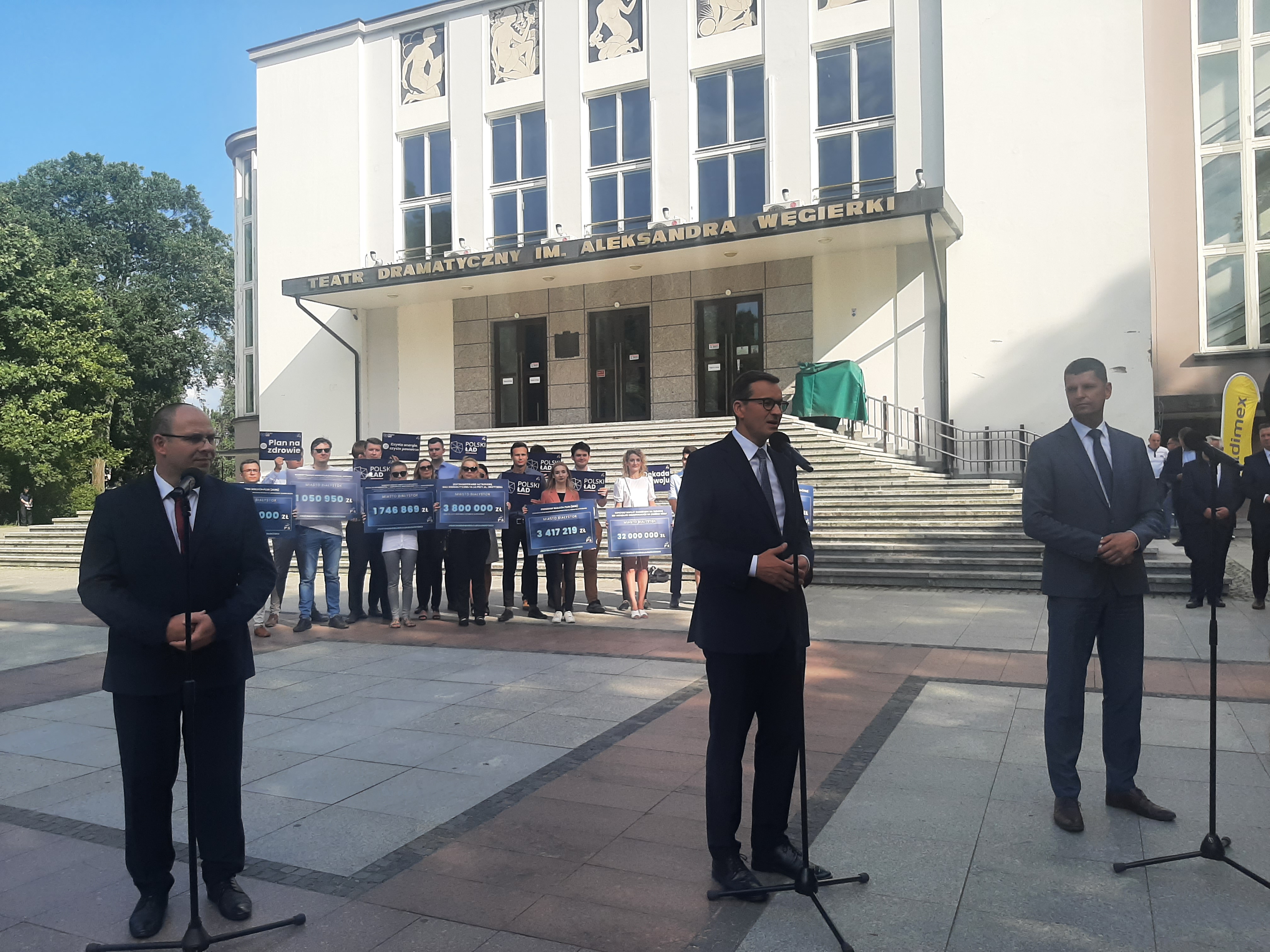 Na pierwszym planie, od lewej stoi dyrektor Teatru Dramatycznego, premier Polski, poseł Dariusz Piątkowski. Stoją na tle Teatru Dramatycznego w Białymstoku. W oddali są osoby trzymające tabliczki z napisami Polski Ład
