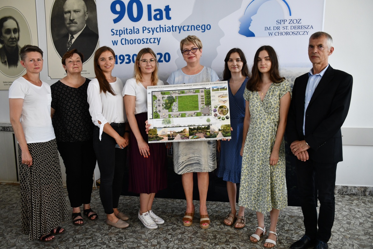 Na ściance z napisem 90. lat Szpitala w Choroszczy stoją m.in. studentki a pośrodku nich dyrektor szpitala, która trzyma w rękach projekt ogrodu przyszpitalnego