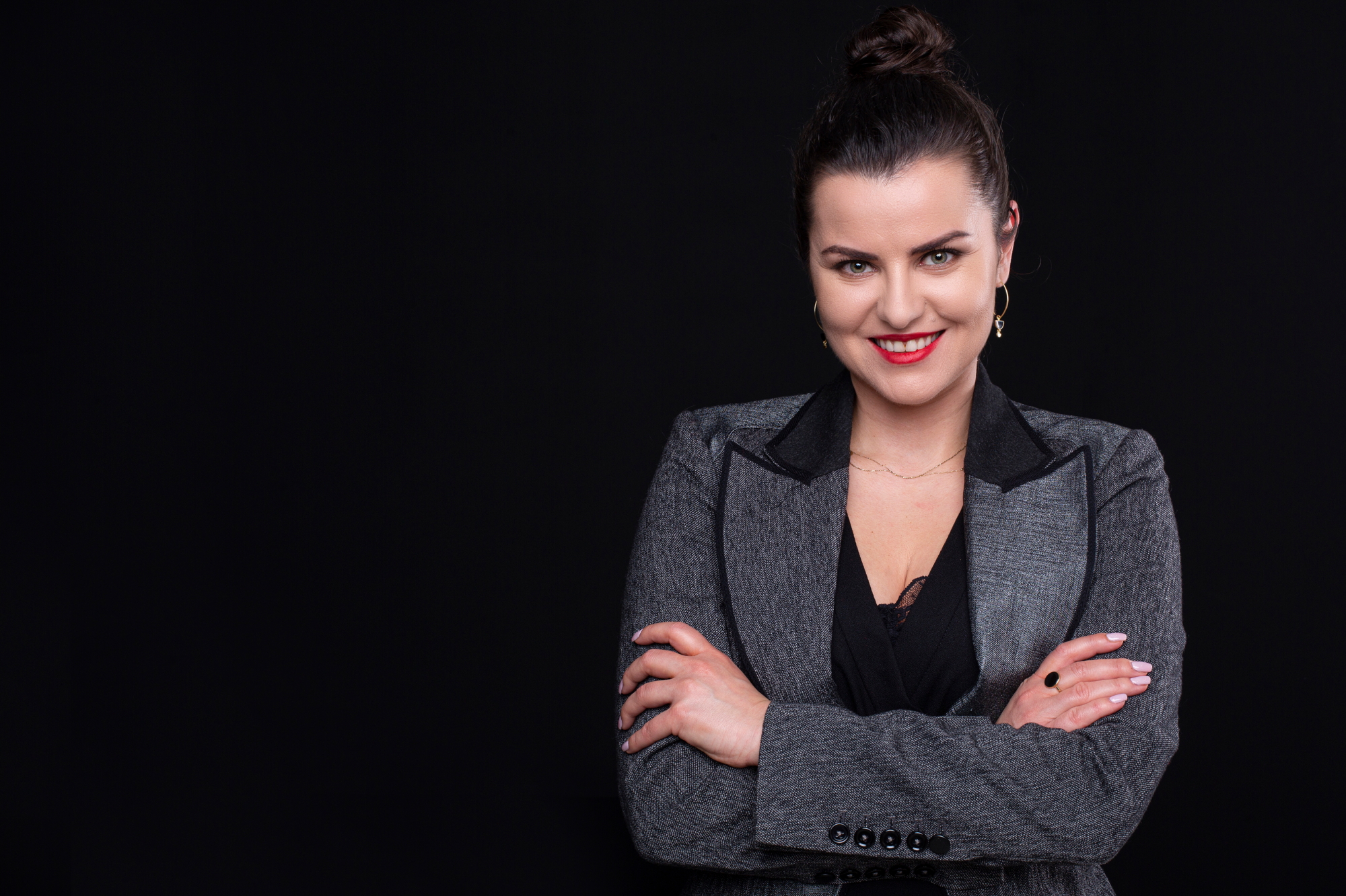 Na czarnym tle stoi uśmiechnięta kobieta, ma związane ciemne włosy, czerwone usta.. Ubrana jest w szarą marynarkę i czarną koszulkę pod spodem