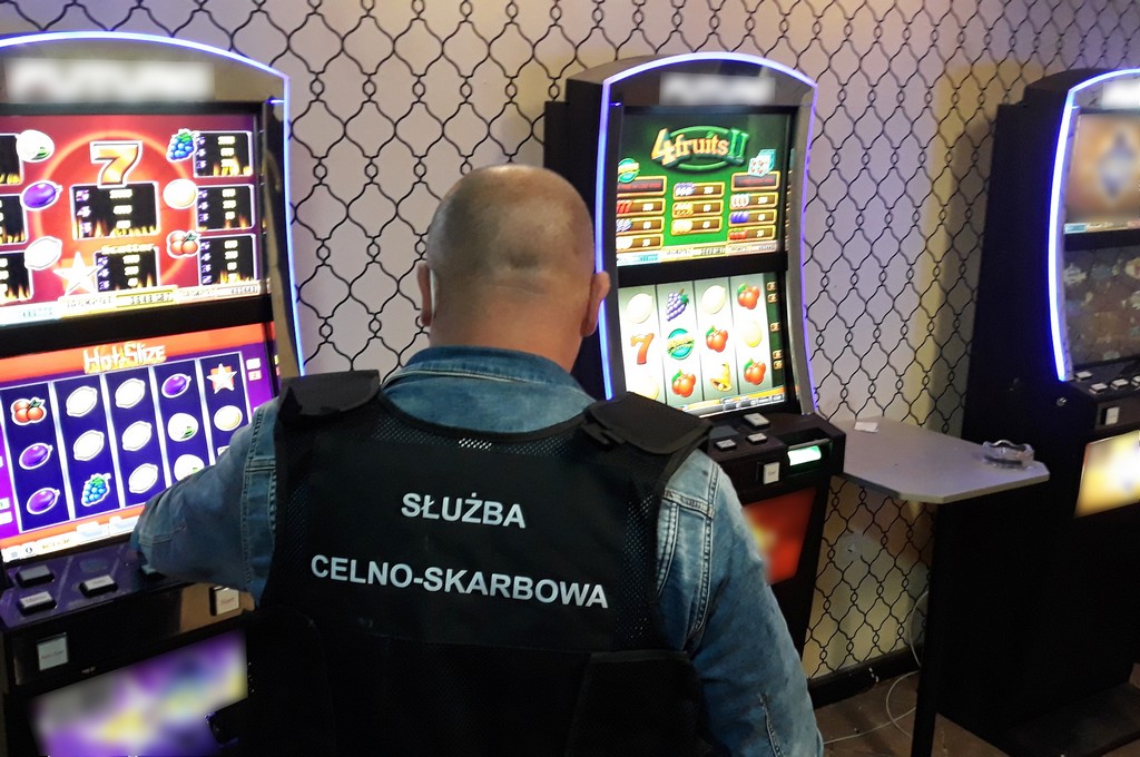 Pod ścianą stoją trzy automaty do gry, przy jednym stoi funkcjonariusz w czarnej kamizelce, na której jest napisane: Służba Celno - Skarbowa