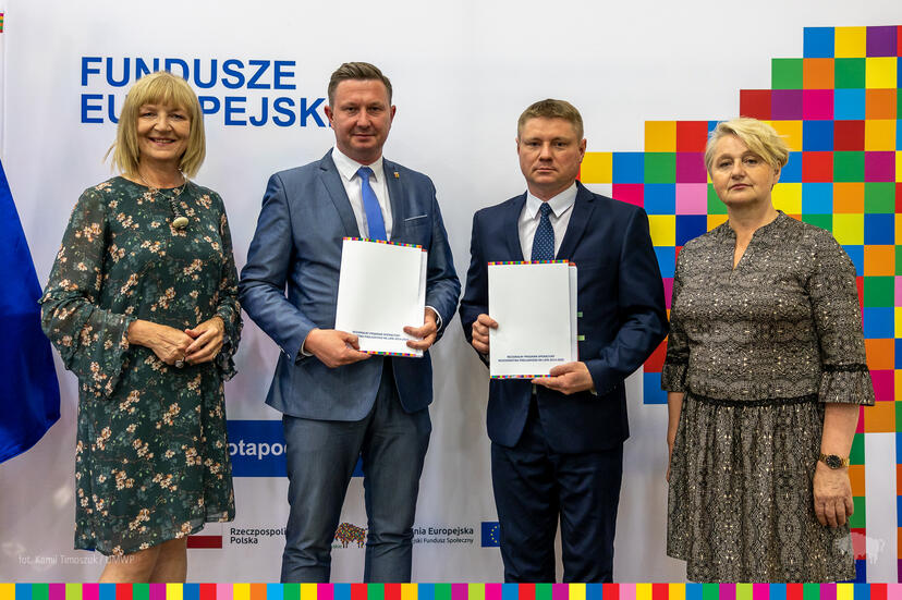 Cztery osoby, od lewej kobieta, po środku dwóch mężczyzn, z prawej kobieta stoją na ściance funduszy europejskich. Mężczyźni trzymają podpisane umowy