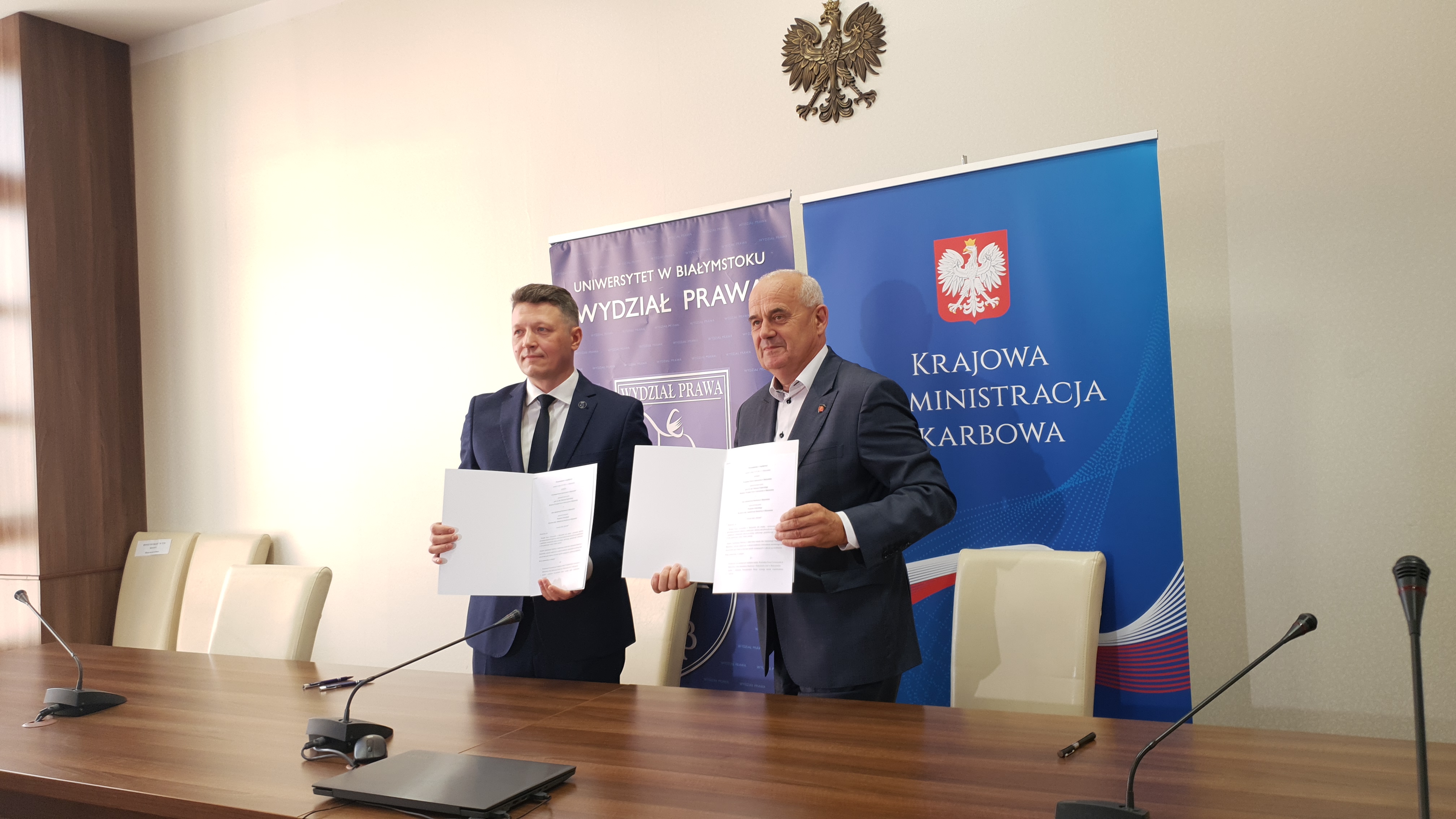 Dwaj mężczyźni stoją przy biurku, w rękach trzymają egzemplarze umów