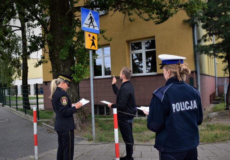 Strażniczka Miejska, Policjantka i urzędnik stoją w okolicy szkoły. Kobiety notują,. mężczyzna przygląda się znakowi drogowemu