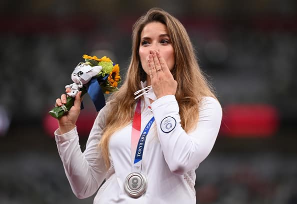 Oszczepniczka Maria Andrejczyk stoi w białej bluzie, w prawej dłoni trzyma maskotkę, lewą ma przytkniętą do ust, na szyi lekkoatletki wisi medal