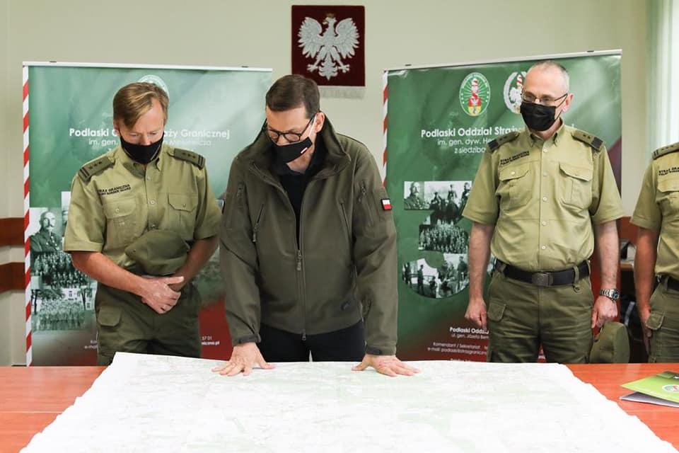 Premier Mateusz Morawicki pochyla się nad stołem, na którym leży mapa. Po jego obu stronach stoją funkcjonariusze Straż Graniczne. Za nimi, stoją standy z napisem: Straż Graniczna