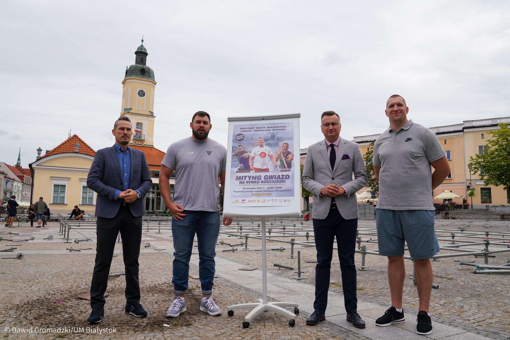 Czterech mężczyzn stoi na Rynku Kościuszki w Białymstoku, między nimi tablica z plakatem informującym o imprezie lekkoatletycznej. W tle widać ratusz