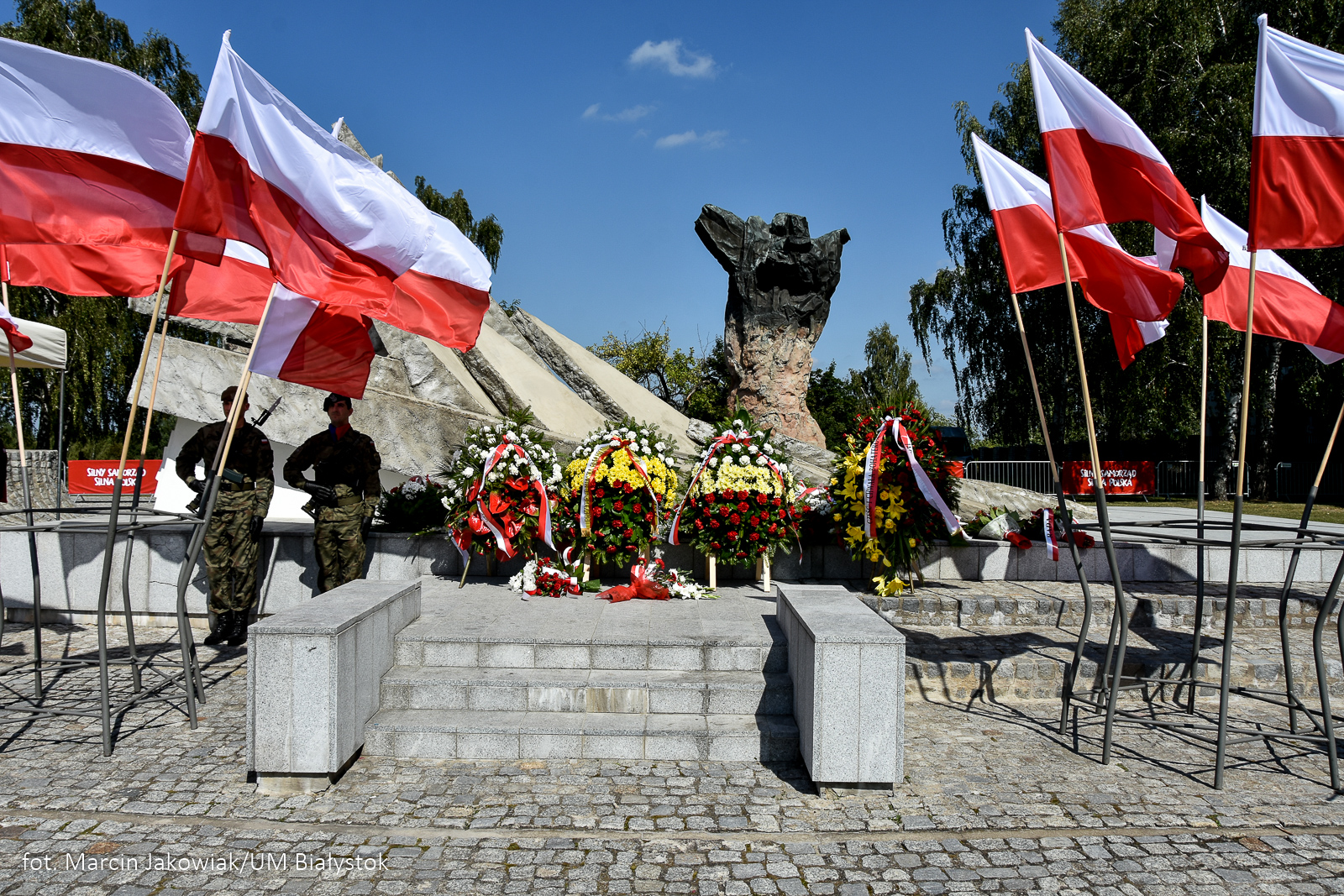 Dwaj żołnierze stoją przy pomniku obrońców miasta, przed pomnikiem stoją wieńce kwiatów, na wietrze trzepoczą biało-czerwone flagi