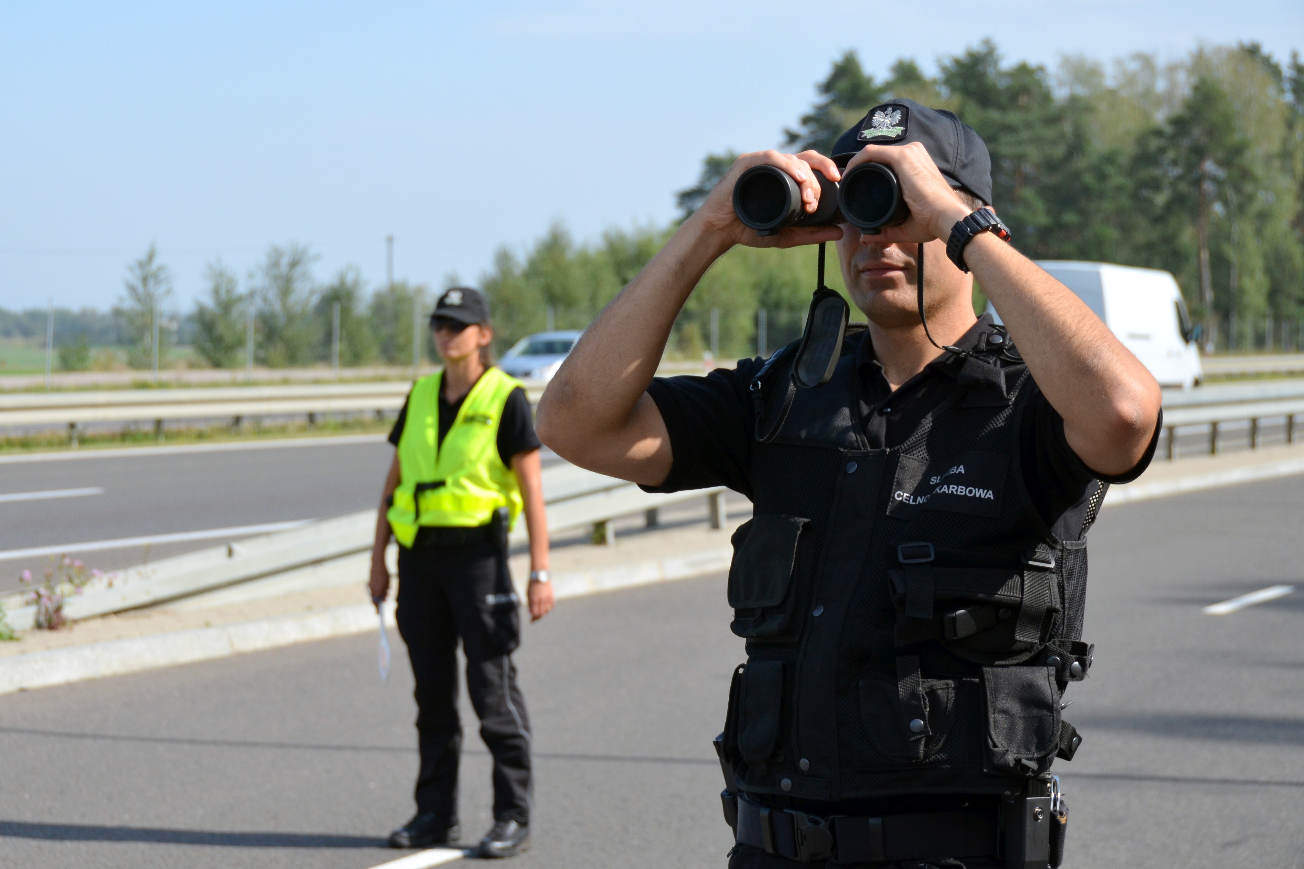 Na pierwszym planie funkcjonariusz z lornetką podczas kontroli drogowej. W tle funkcjonariuszka, pas drogowy i samochody osobowe.